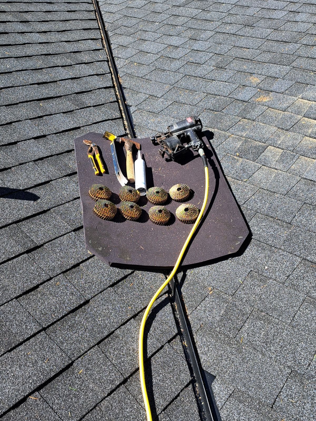 Tools and equipment for roof work laid out on a black surface on a shingled roof, including a power drill, gloves, and various grinding or polishing discs.