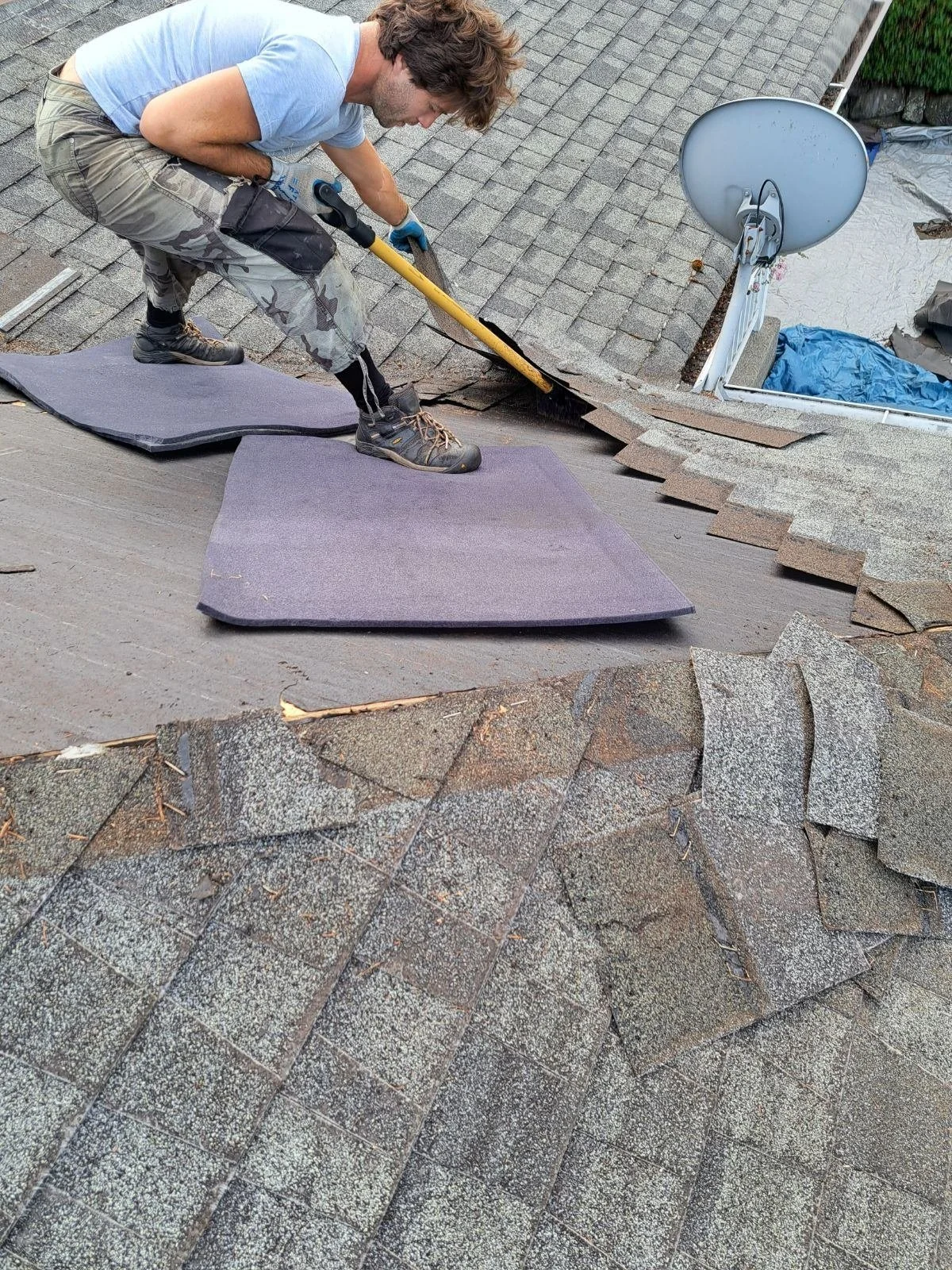A person working on a roof, lifting and removing shingles with a crowbar, with roofing tools and materials nearby.
