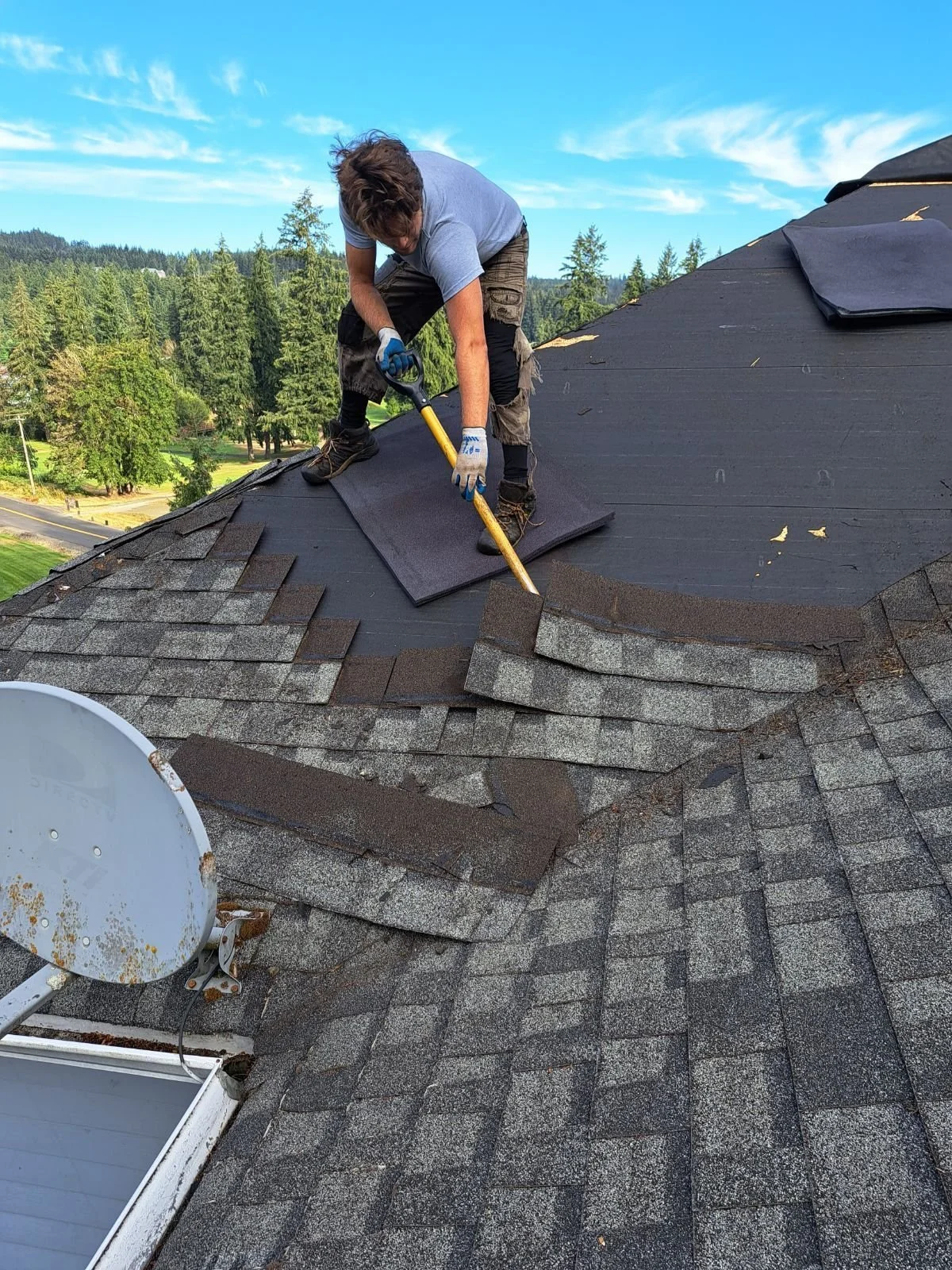 A man working on a rooftop, installing or repairing roofing shingles on a sunny day with a clear blue sky and green trees in the background.
