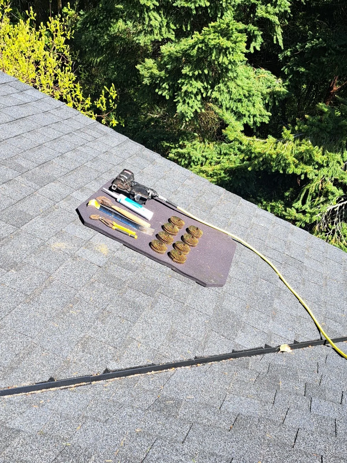 Tools and equipment placed on a black mat on a rooftop, including a cordless drill, screwdrivers, a small flashlight, a tube, screws, and a yellow extension cord, with green trees in the background.