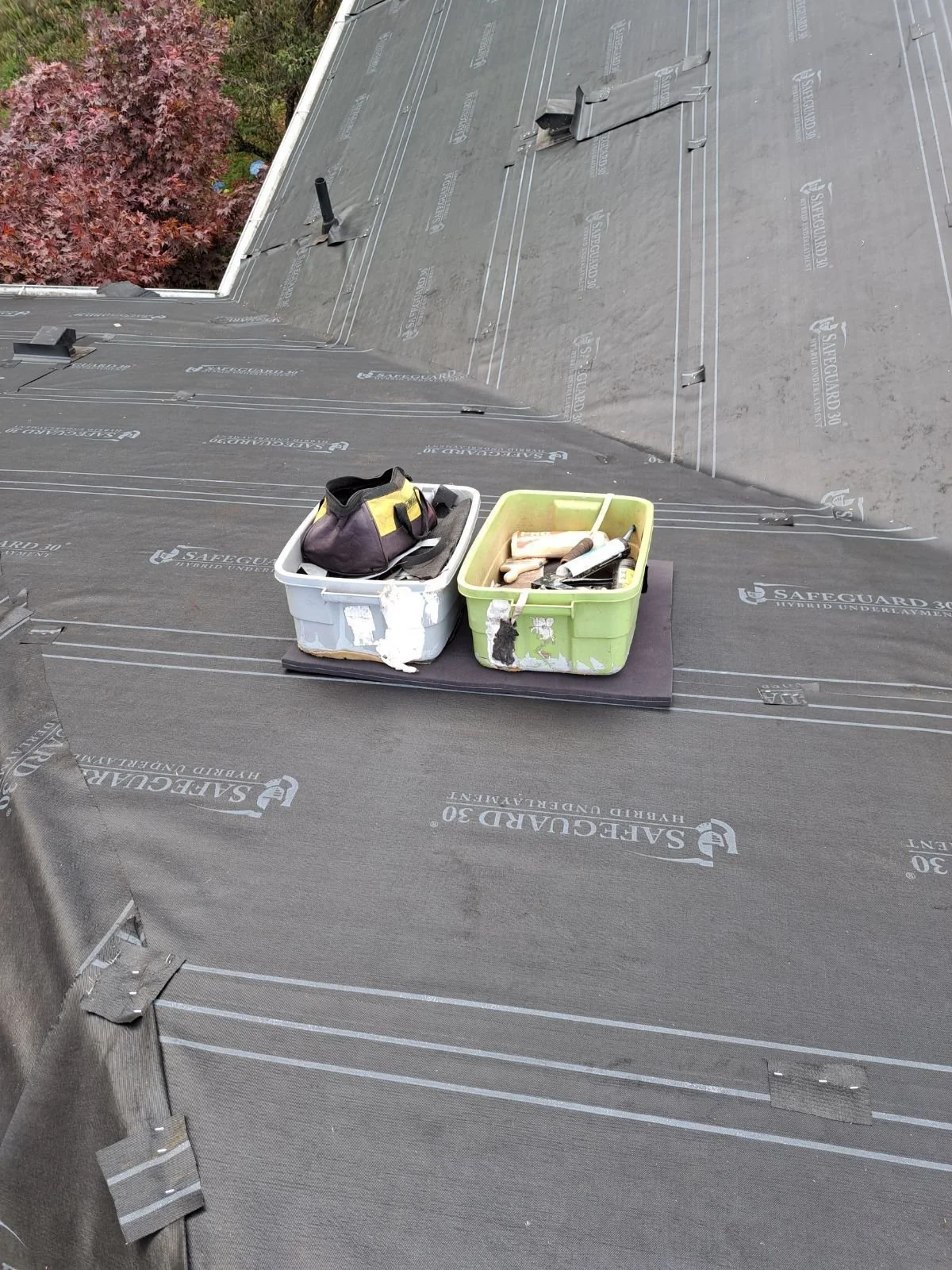 A section of a roof under construction, showing roofing underlayment with tools and a bag on top of two containers placed on black material.