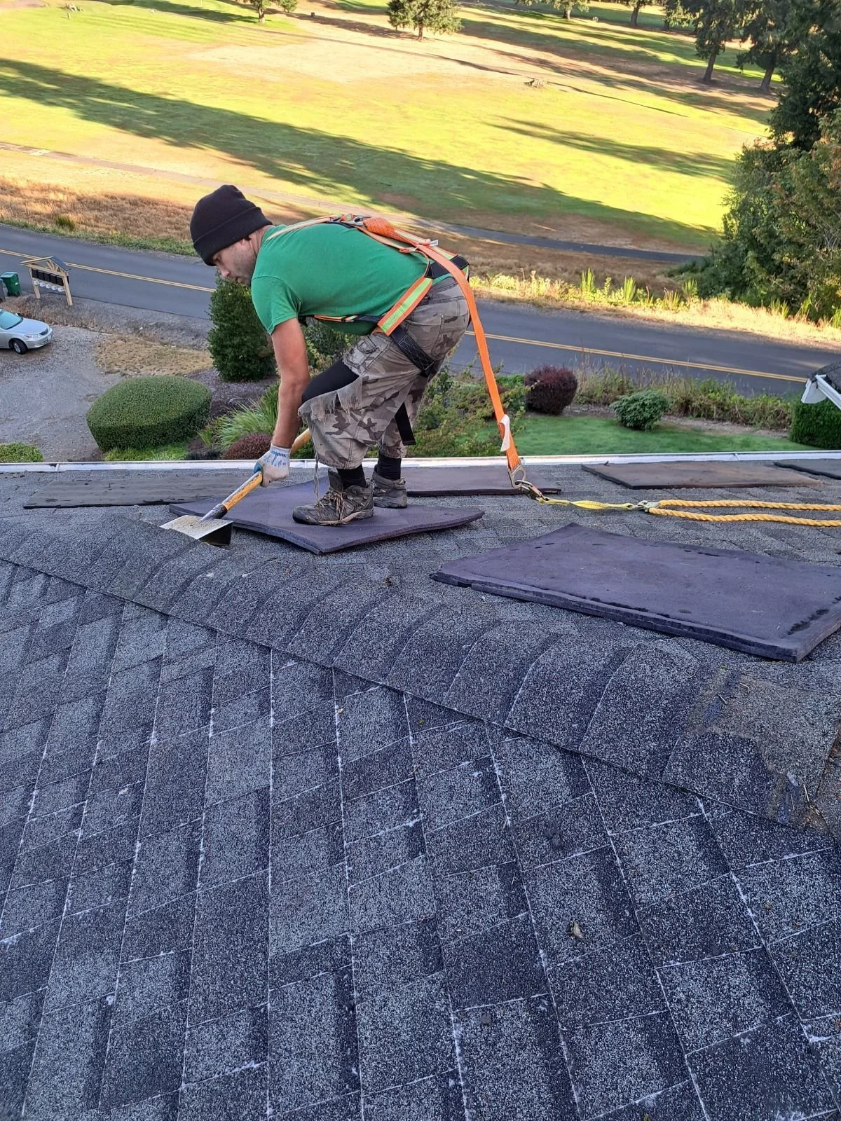A man is working on a roof, lifting and adjusting shingles while standing on a padded safety harness anchored to the roof. There are tools and mats nearby, with trees and a road in the background.