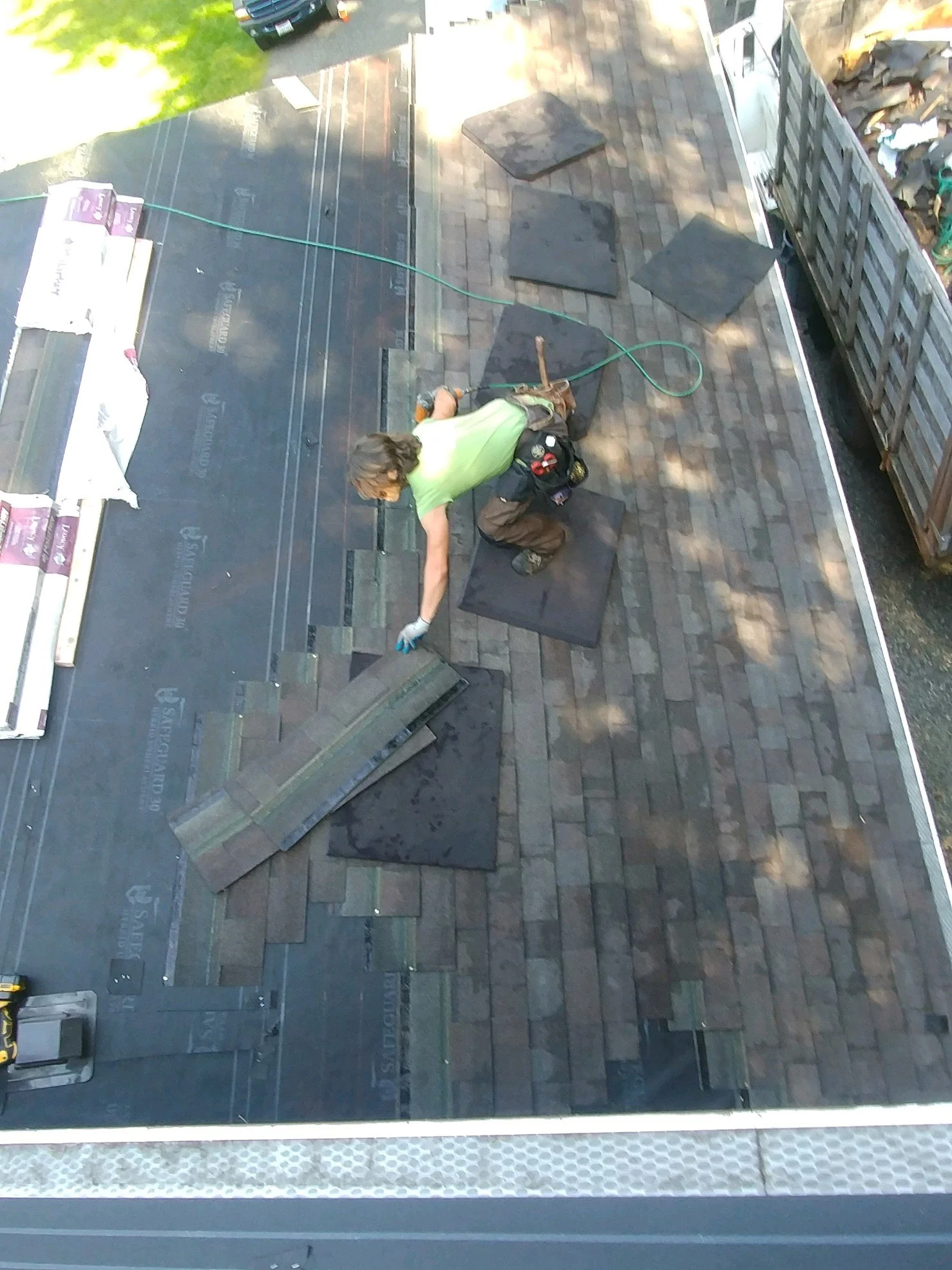 A person working on installing shingles on a roof, kneeling on black padding, with tools nearby, and stacks of shingles ready to be put in place.