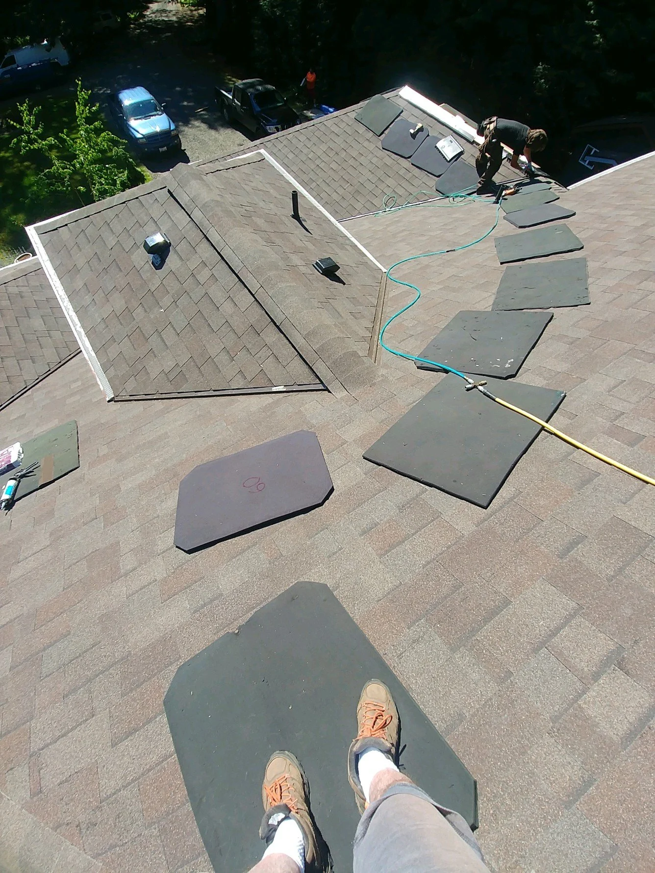 View of a rooftop with shingles and solar panels, worker installing or repairing panels, and a person's feet on a safety harness or platform, with a parking lot and trees below.