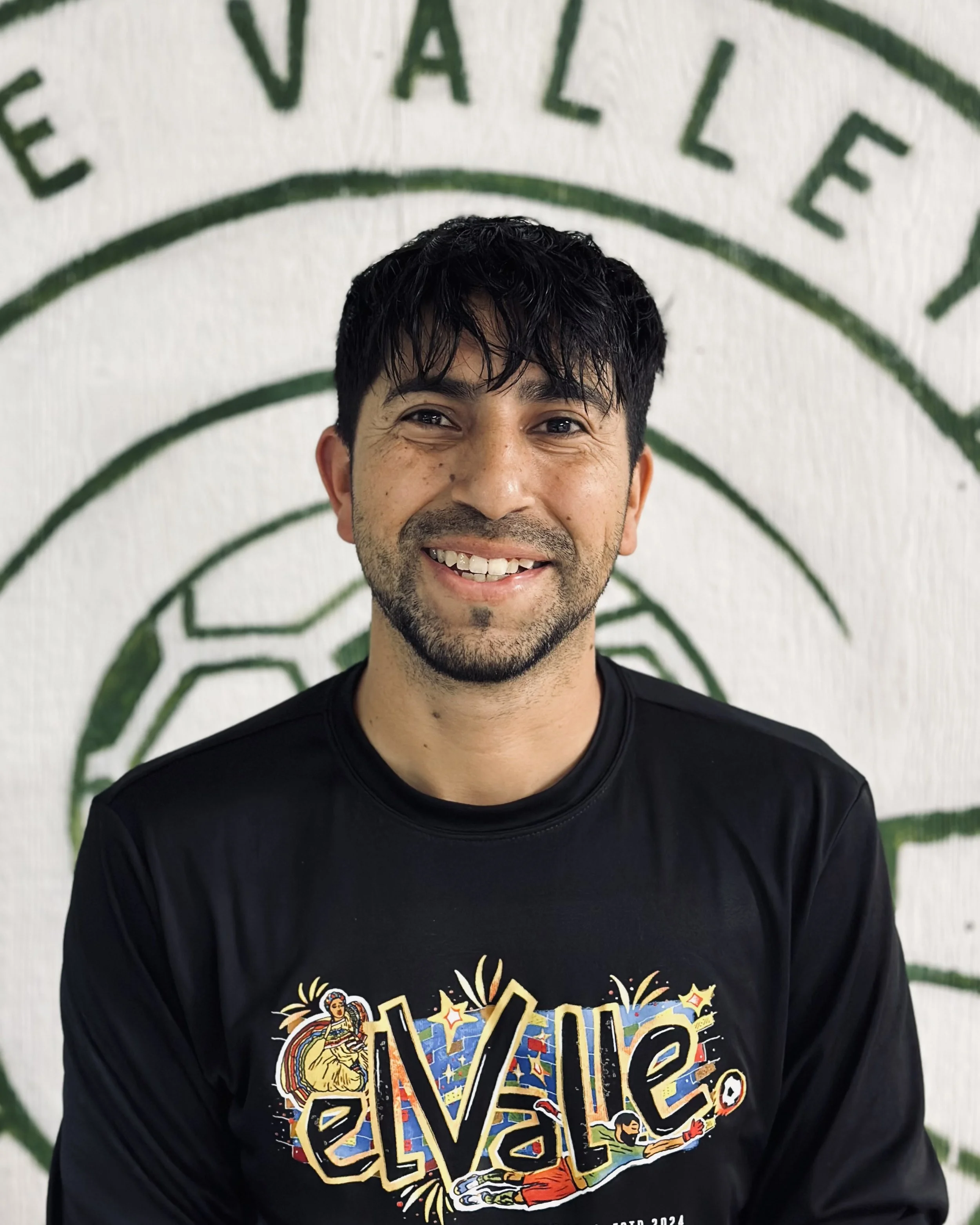 Smiling man with dark hair, wearing a black shirt with colorful text and illustrations, standing in front of a wall with green text reading "Valley".