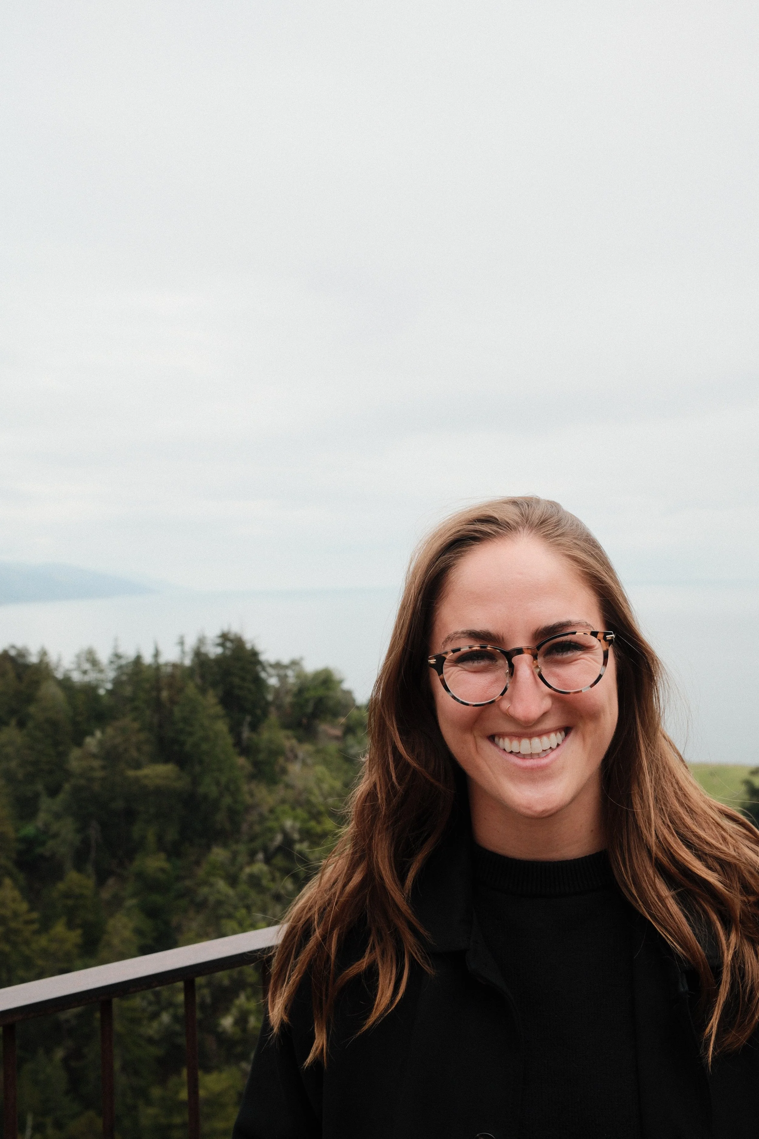 Smiling woman with glasses and long hair outdoors, with a green hilly landscape and cloudy sky in the background.