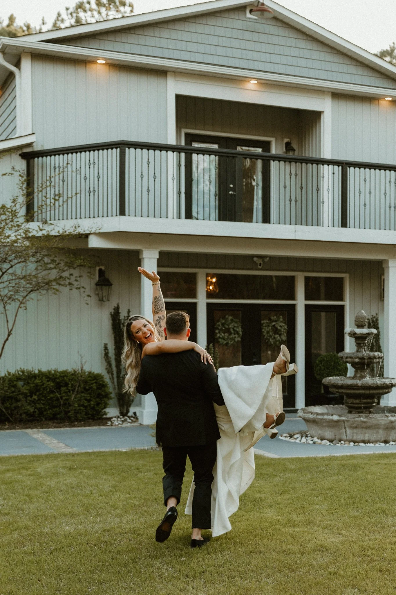 A newlywed couple dancing on the lawn in front of a large house with a balcony and outdoor fountain, during the evening.