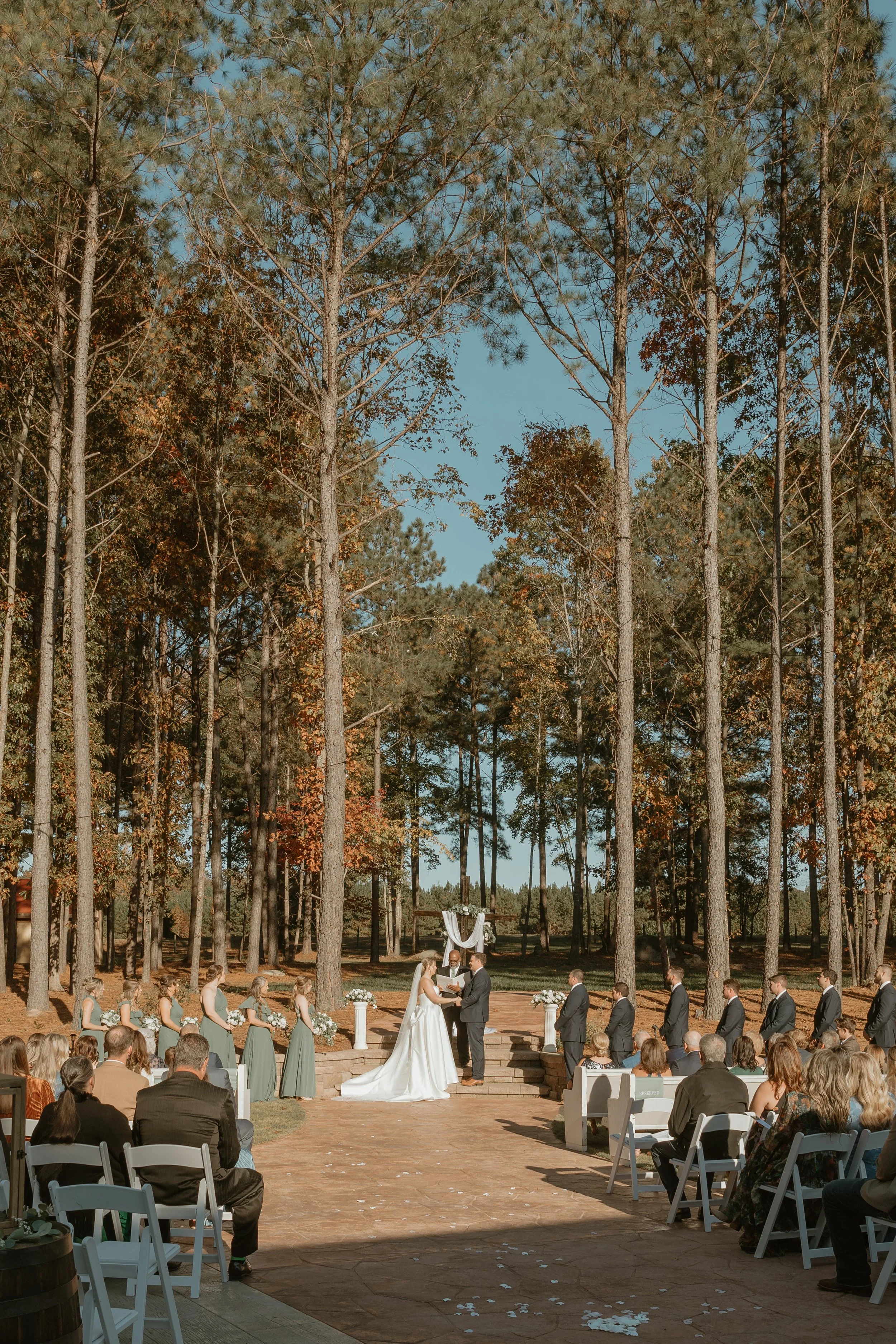 A wedding ceremony taking place outdoors in a wooded area, with the bride and groom standing on steps exchanging vows, surrounded by wedding party and seated guests.
