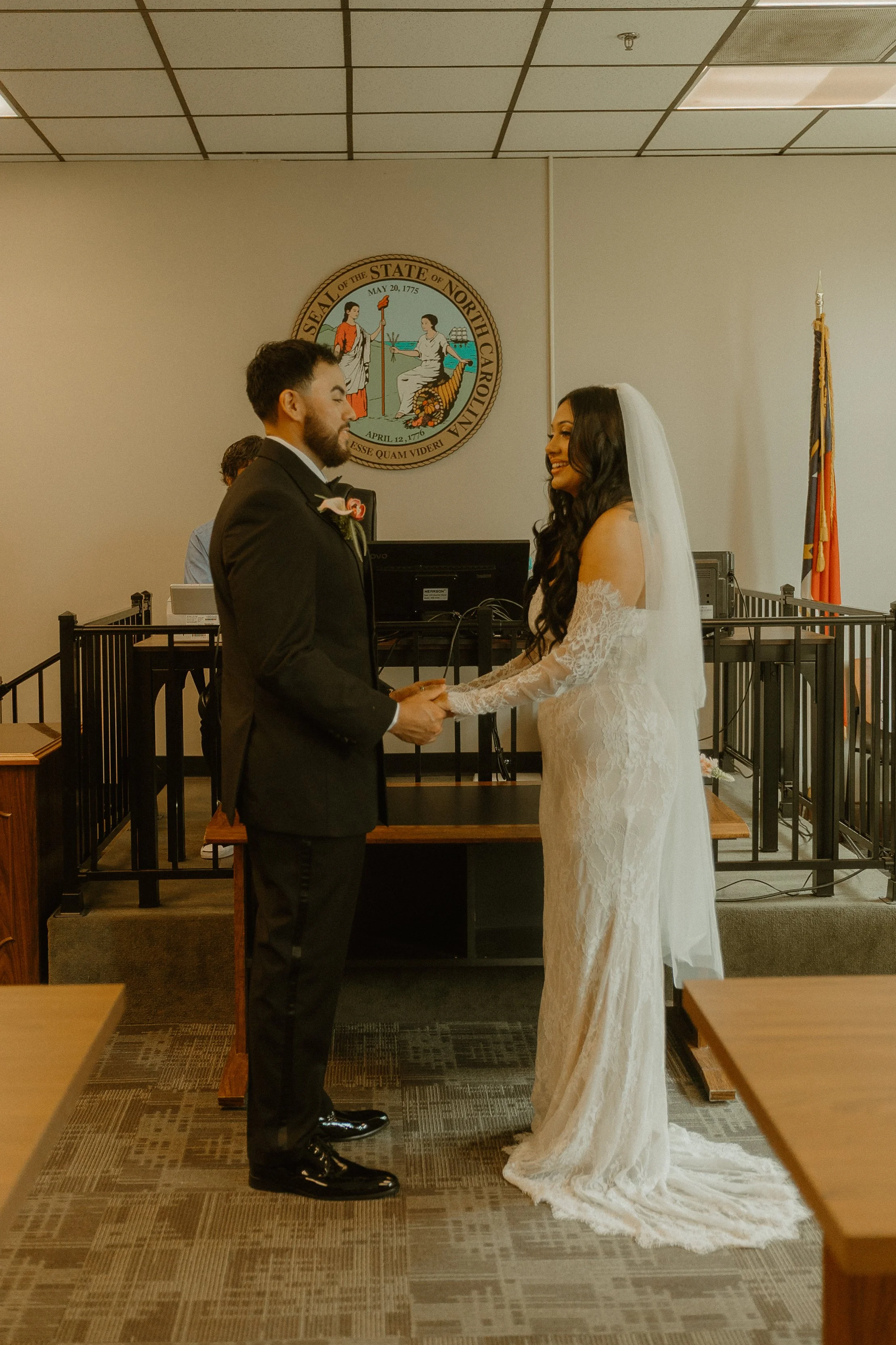 A bride and groom holding hands and smiling at each other during their wedding ceremony in a government building with a North Carolina seal on the wall and an American flag in the background.
