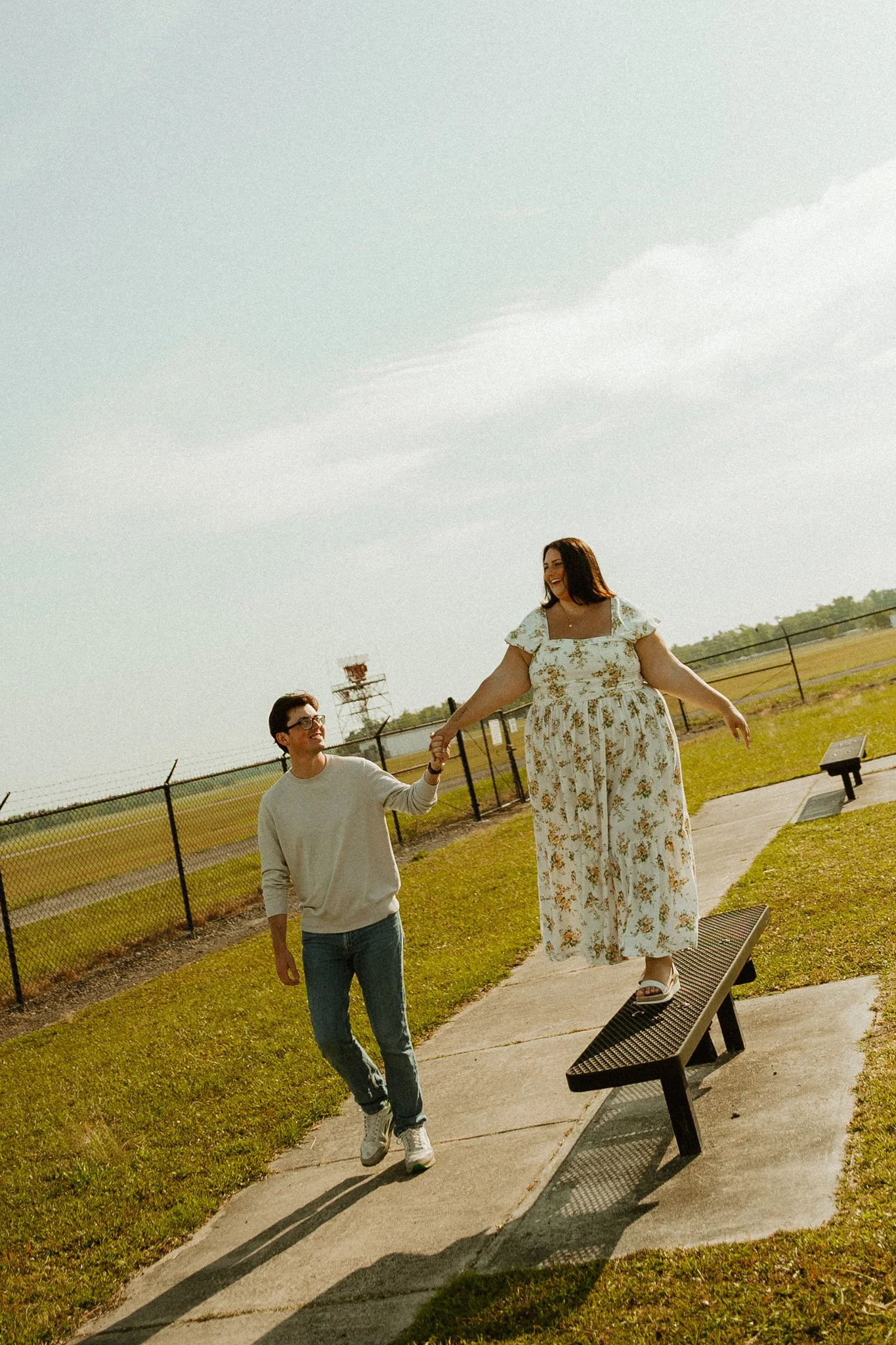 A woman in a floral dress standing on a bench holding hands with a man in casual clothing, walking outdoors near a fence and a grassy area under a cloudy sky.