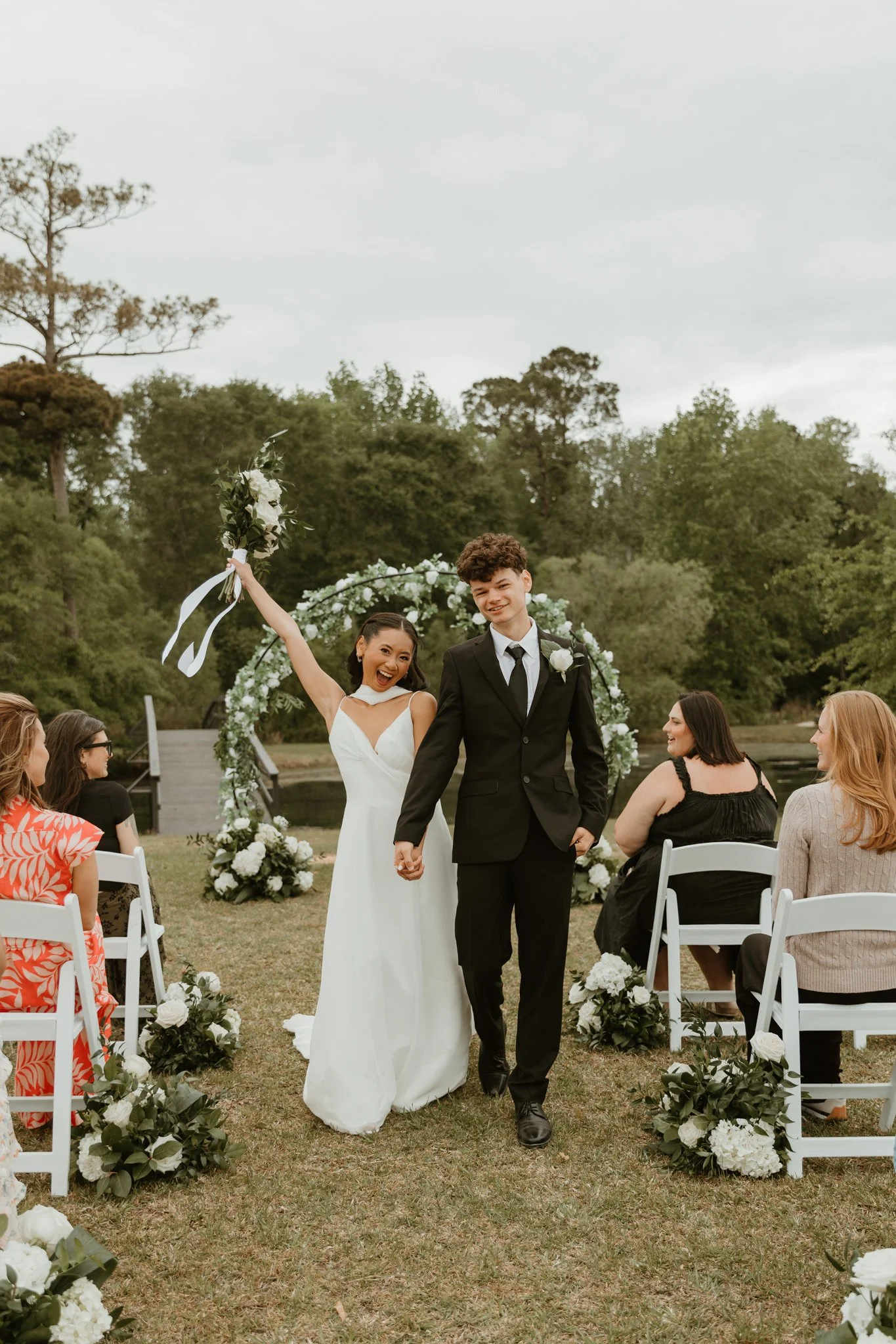 A bride and groom walking hand in hand near a body of water during sunset, with trees and a small orange-leaved tree in the background.