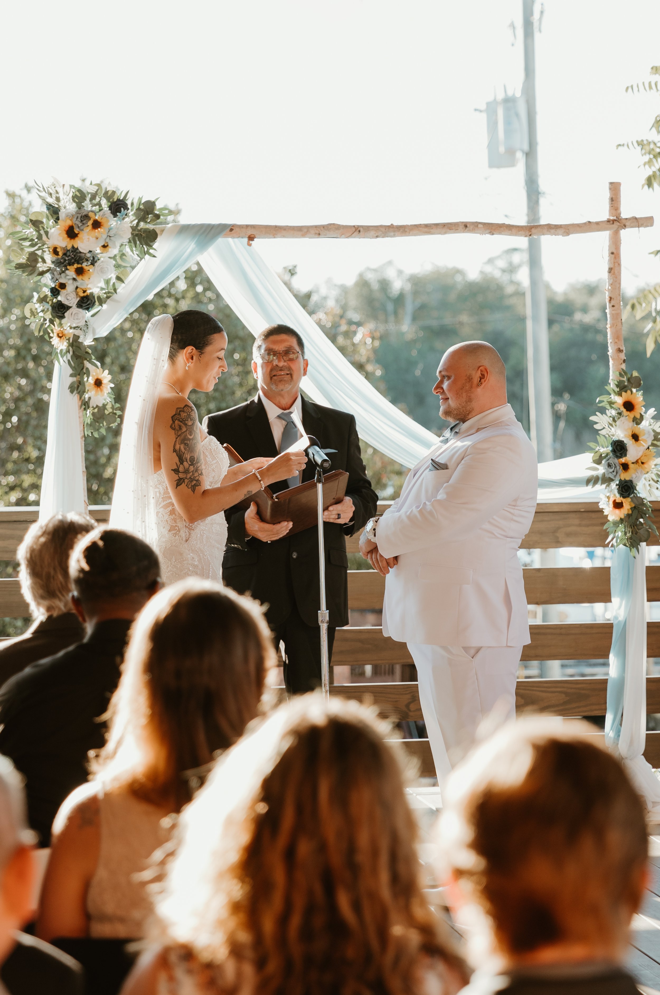 A wedding ceremony with a bride in a white lace dress and veil, a groom in a white tuxedo, and an officiant in a black suit, performing the vows under a wooden arch decorated with white fabric and sunflowers, with guests seated and watching.