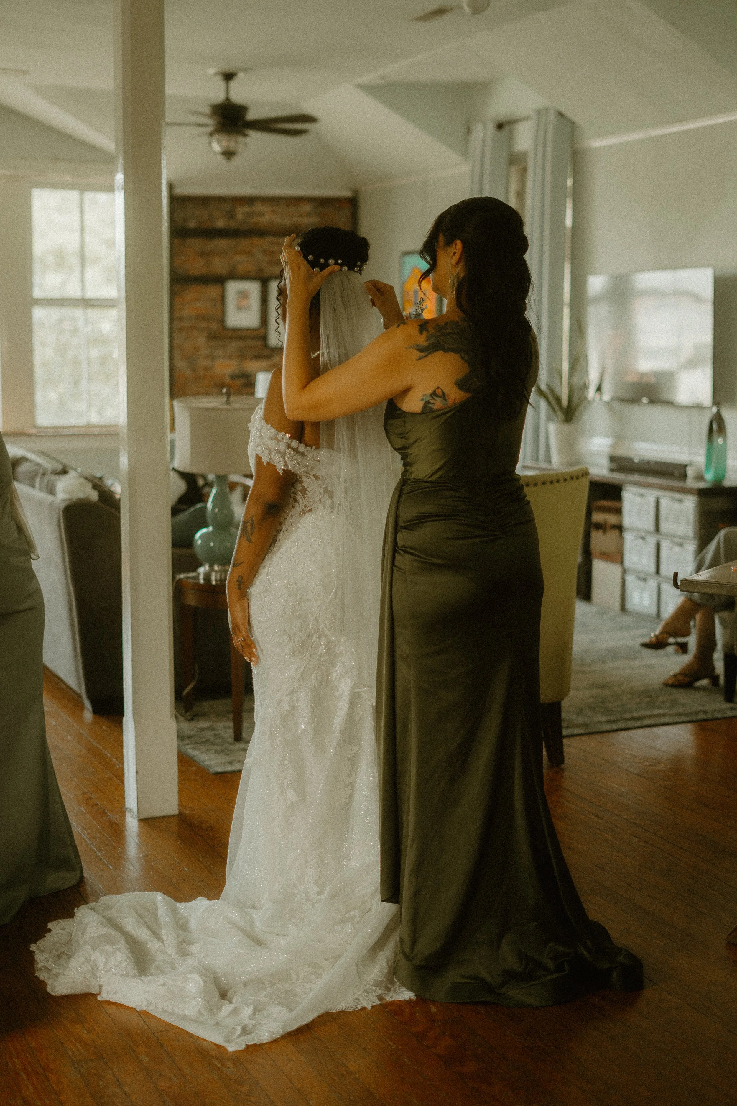 A woman in a black dress is helping a bride adjust her dress and veil in a room with a hardwood floor and a mirror, with other people visible in the background.