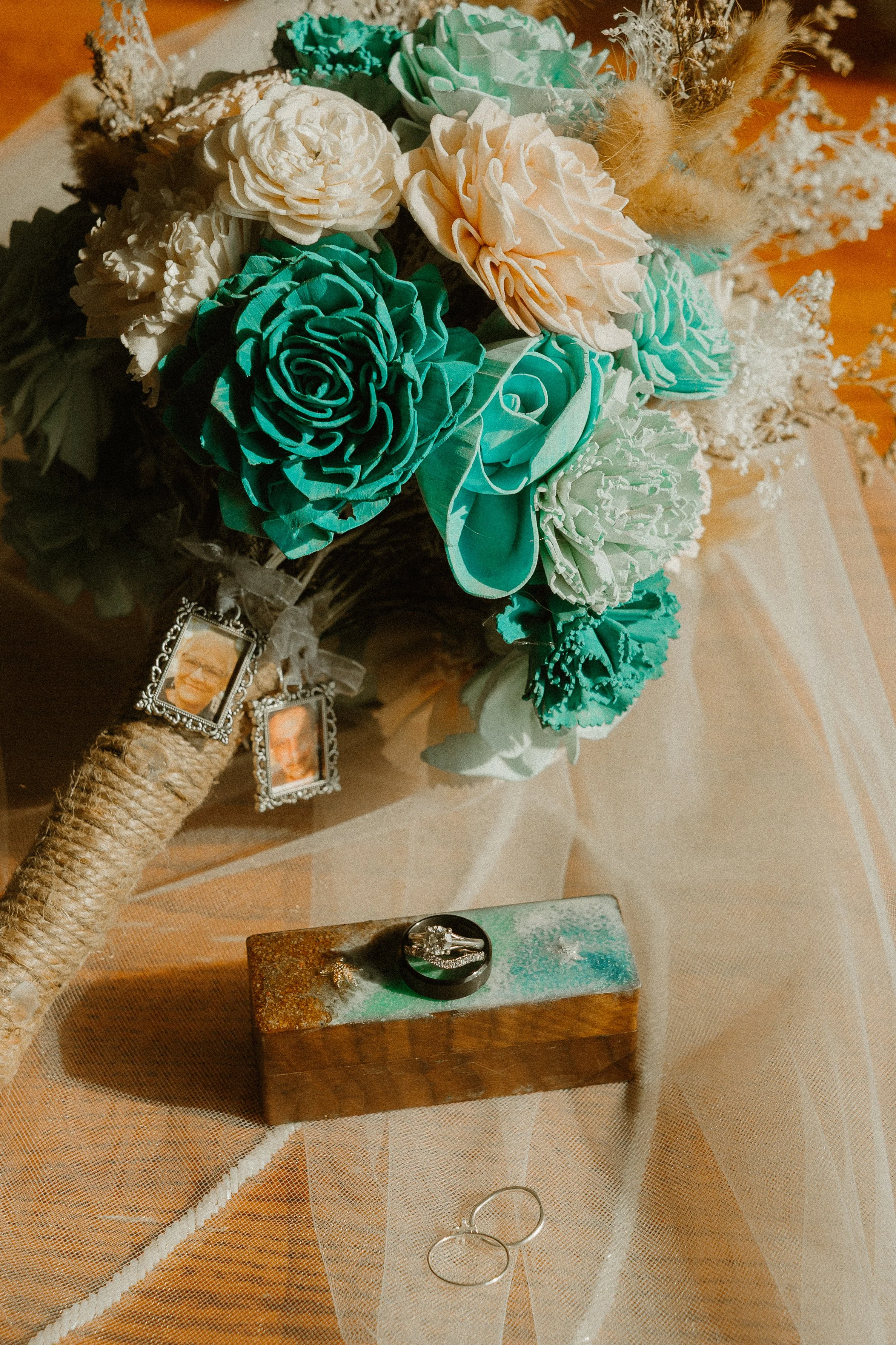 A bouquet of paper flowers in shades of cream, peach, and teal, with greenery and decorative dried plants. The bouquet has picture frames attached, and the background features a wooden table and a ring display with two rings on a small rectangular st