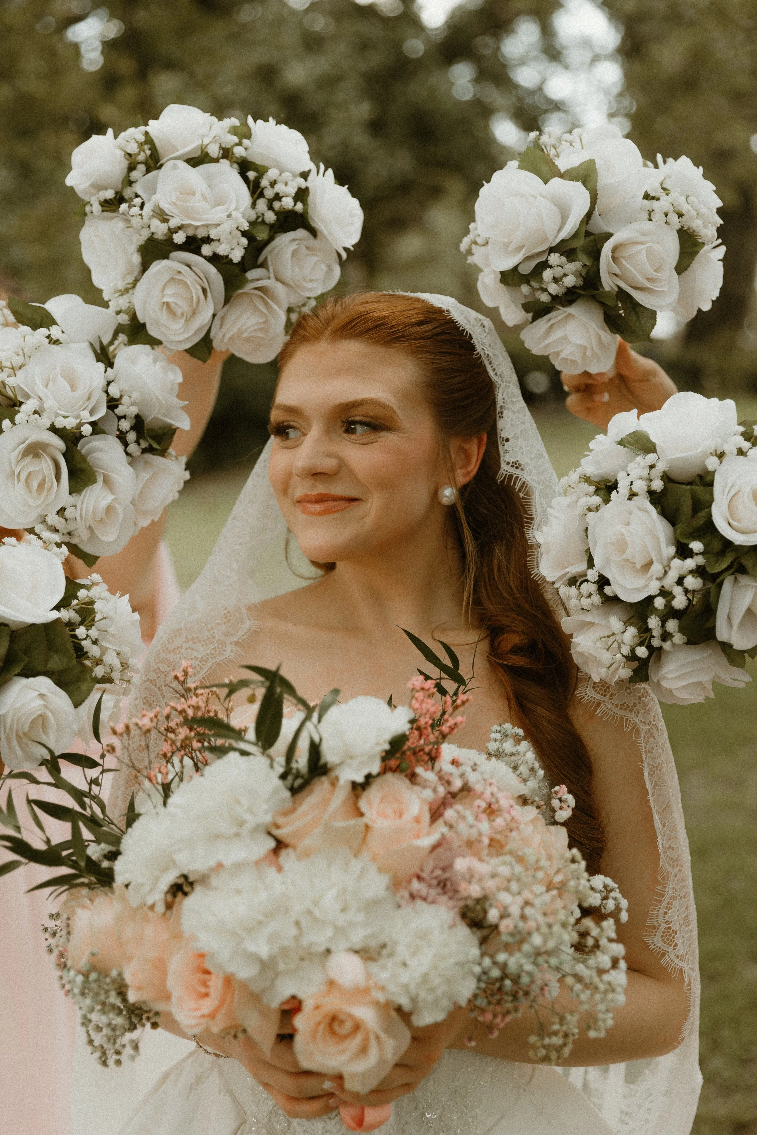 A bride holding a bouquet of pink and white flowers, with her face partially visible. She is wearing a wedding dress and a lace veil, and has auburn hair styled in loose waves.