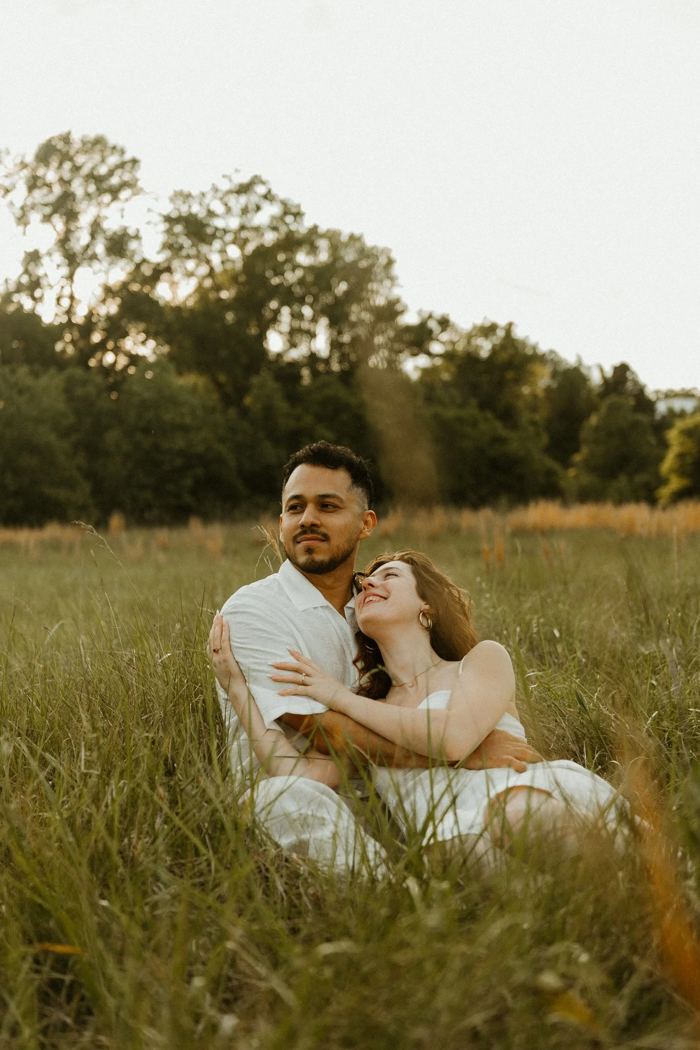 A couple embracing and sitting in tall grass in a field during sunset, with trees in the background.