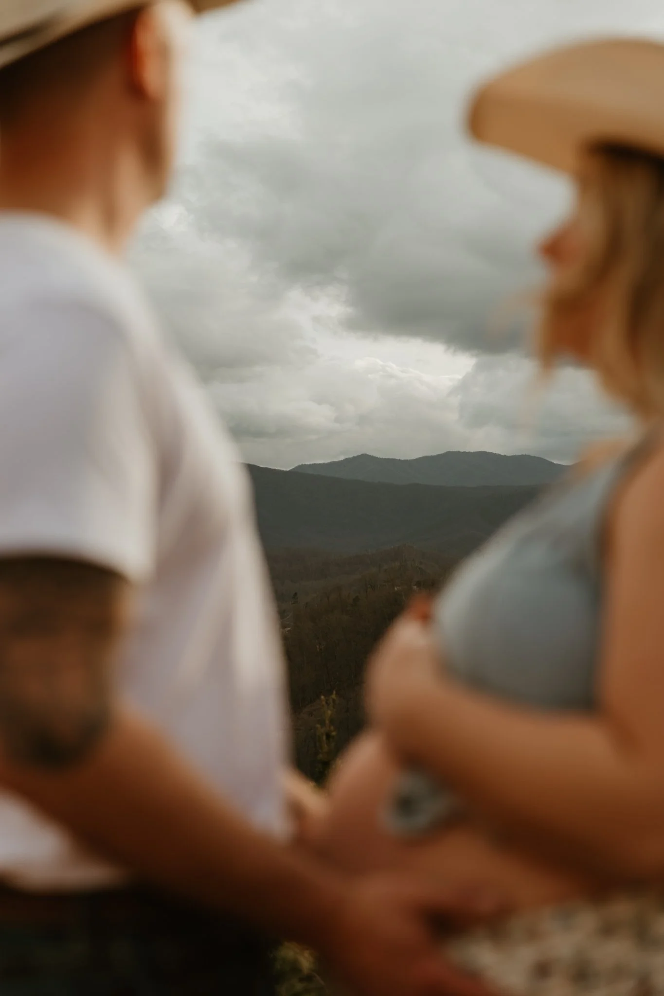 A couple standing outdoors against a mountain landscape with cloudy sky, with the focus on the blurred silhouette of the man and woman.