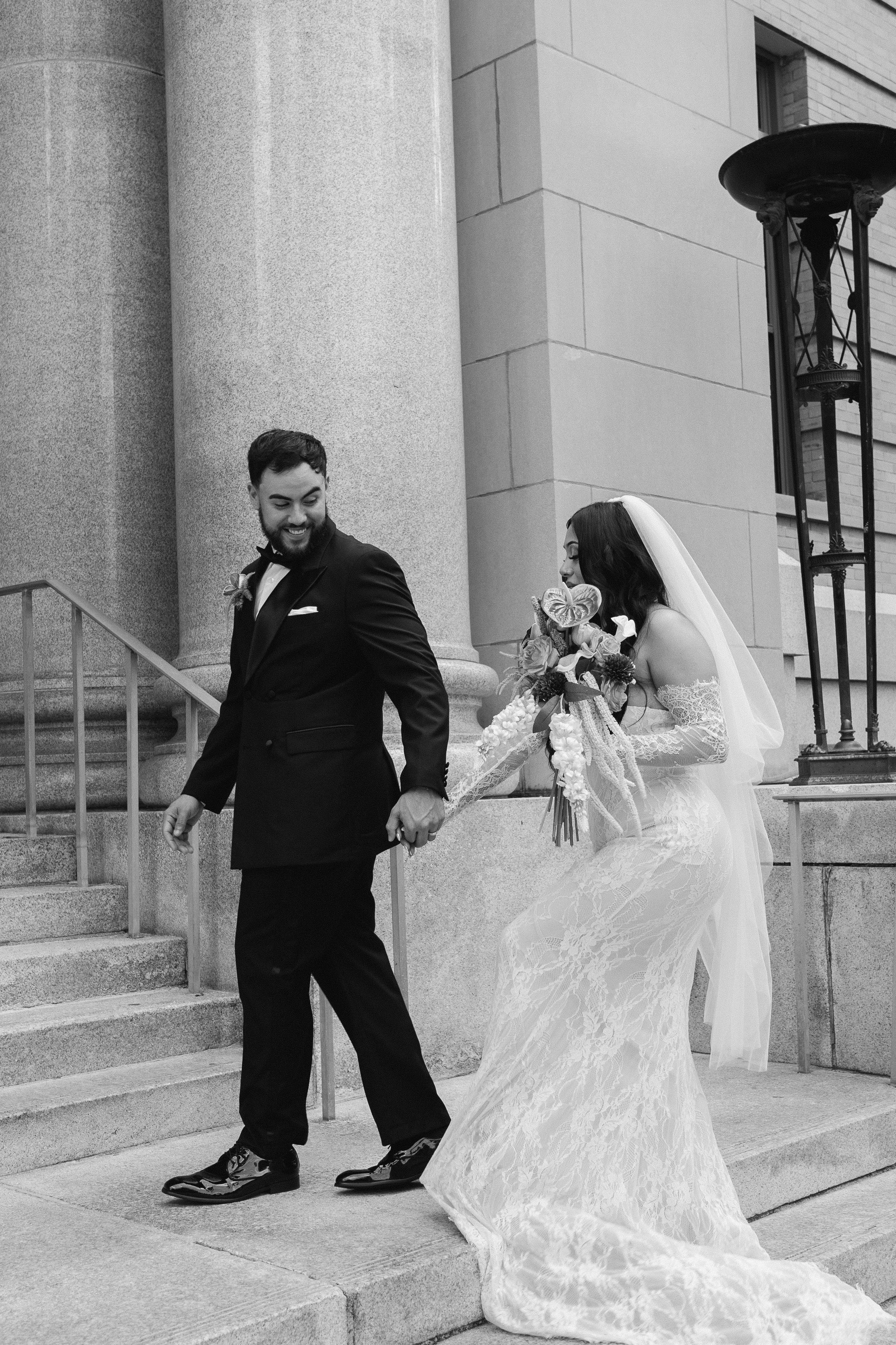 A bride and groom walking down the steps outside a building, holding hands. The groom is wearing a black suit and the bride is in a white wedding dress holding a bouquet.
