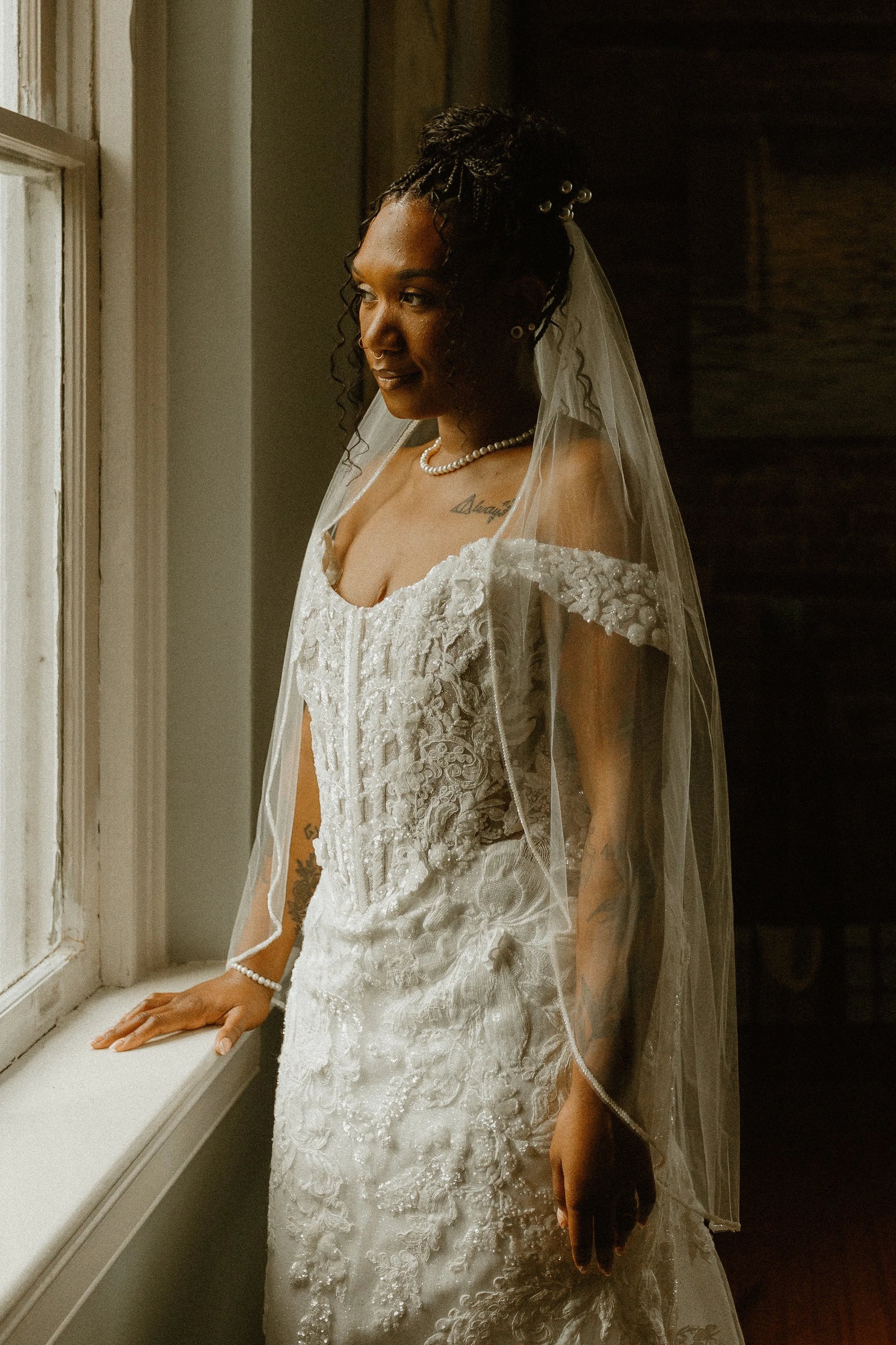 A bride standing by a window in a wedding dress and veil, looking out of the window with her hand on the windowsill, in warm, natural light.