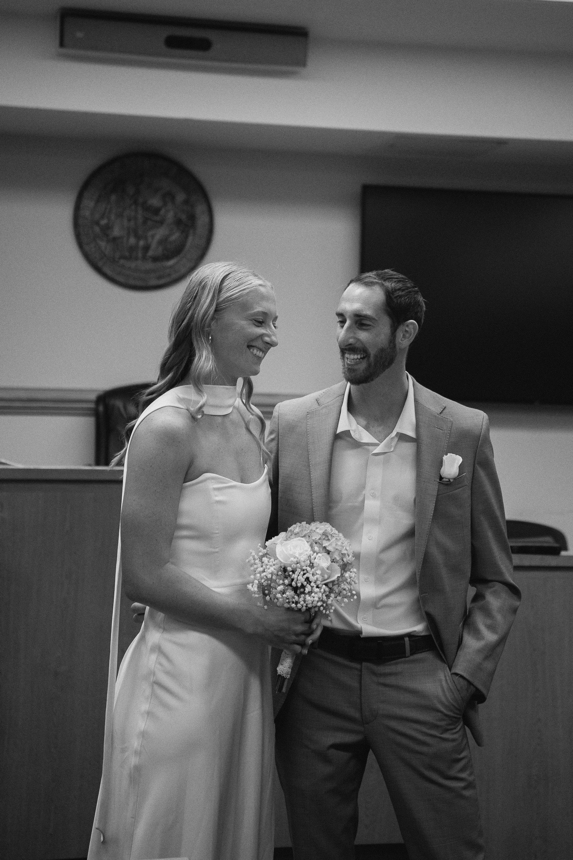 A bride and groom smiling at each other during their wedding ceremony, with the bride holding a bouquet of flowers in a wedding hall.