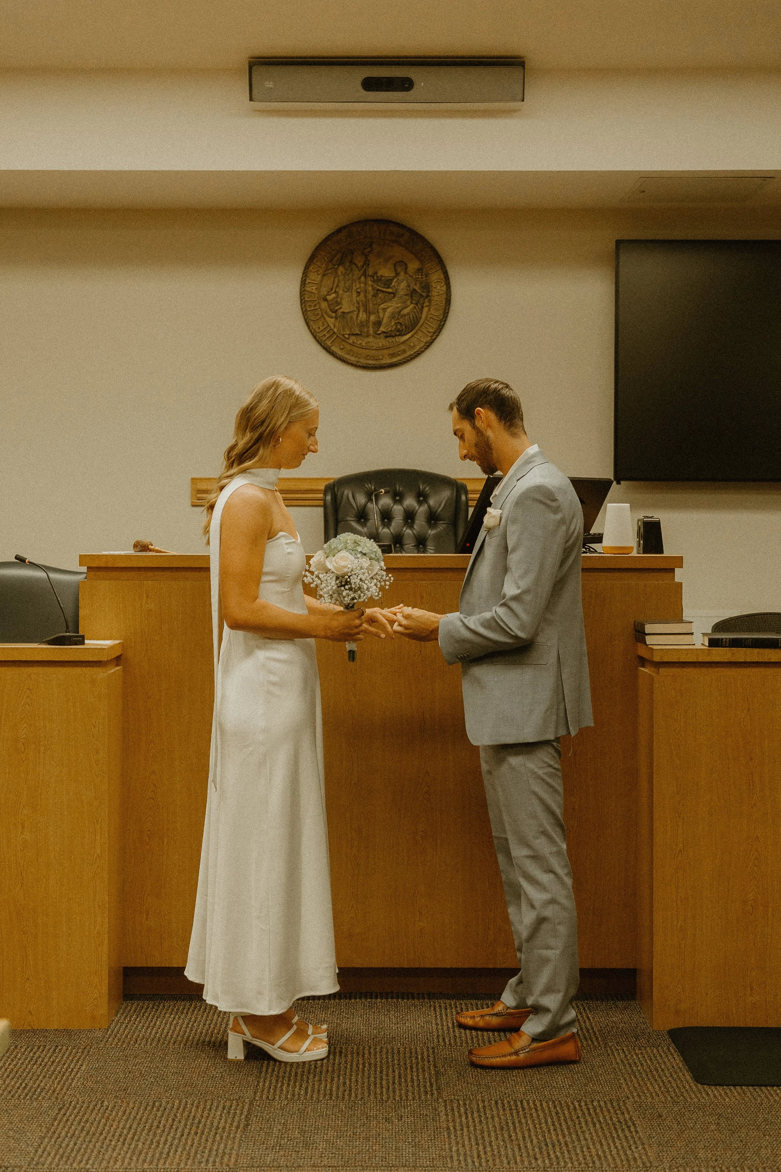 A bride and groom holding hands and looking at each other in a courtroom, with a judge's bench and seal in the background.