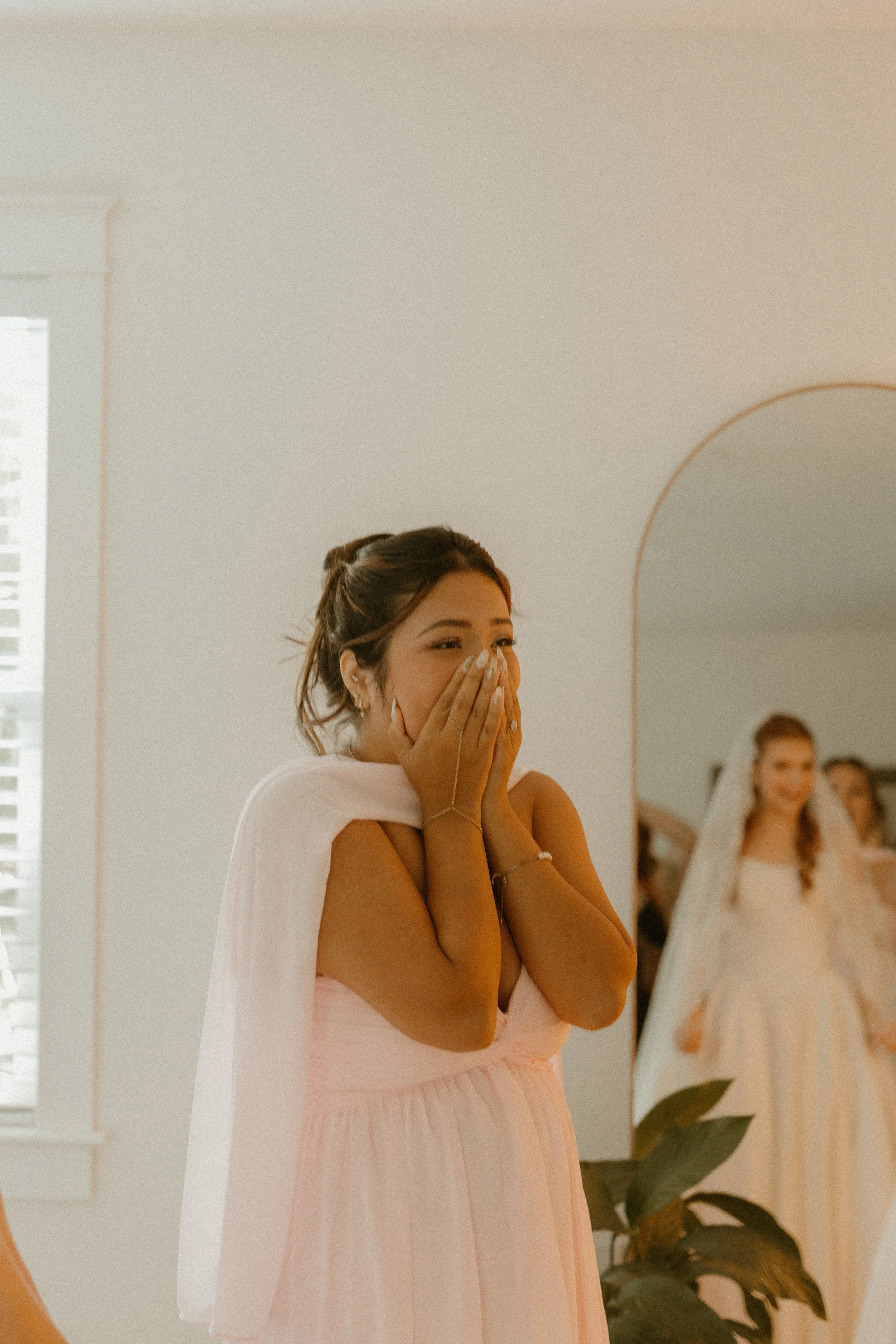 A woman wearing a pink dress is covering her mouth with her hands, appearing emotional or surprised. She is in a room with a mirror and other women, possibly at a wedding or special event.