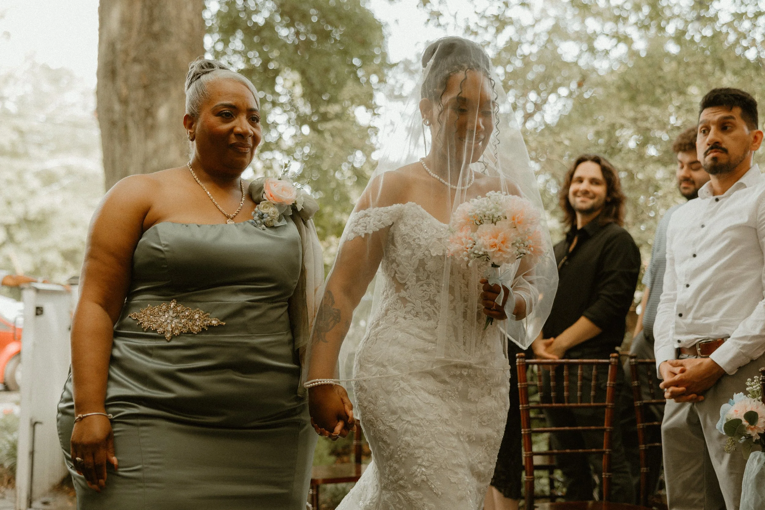 A group of people holding hands during a wedding ceremony outdoors, with a woman in a wedding dress and veil, and others dressed formally, standing in a forested area.