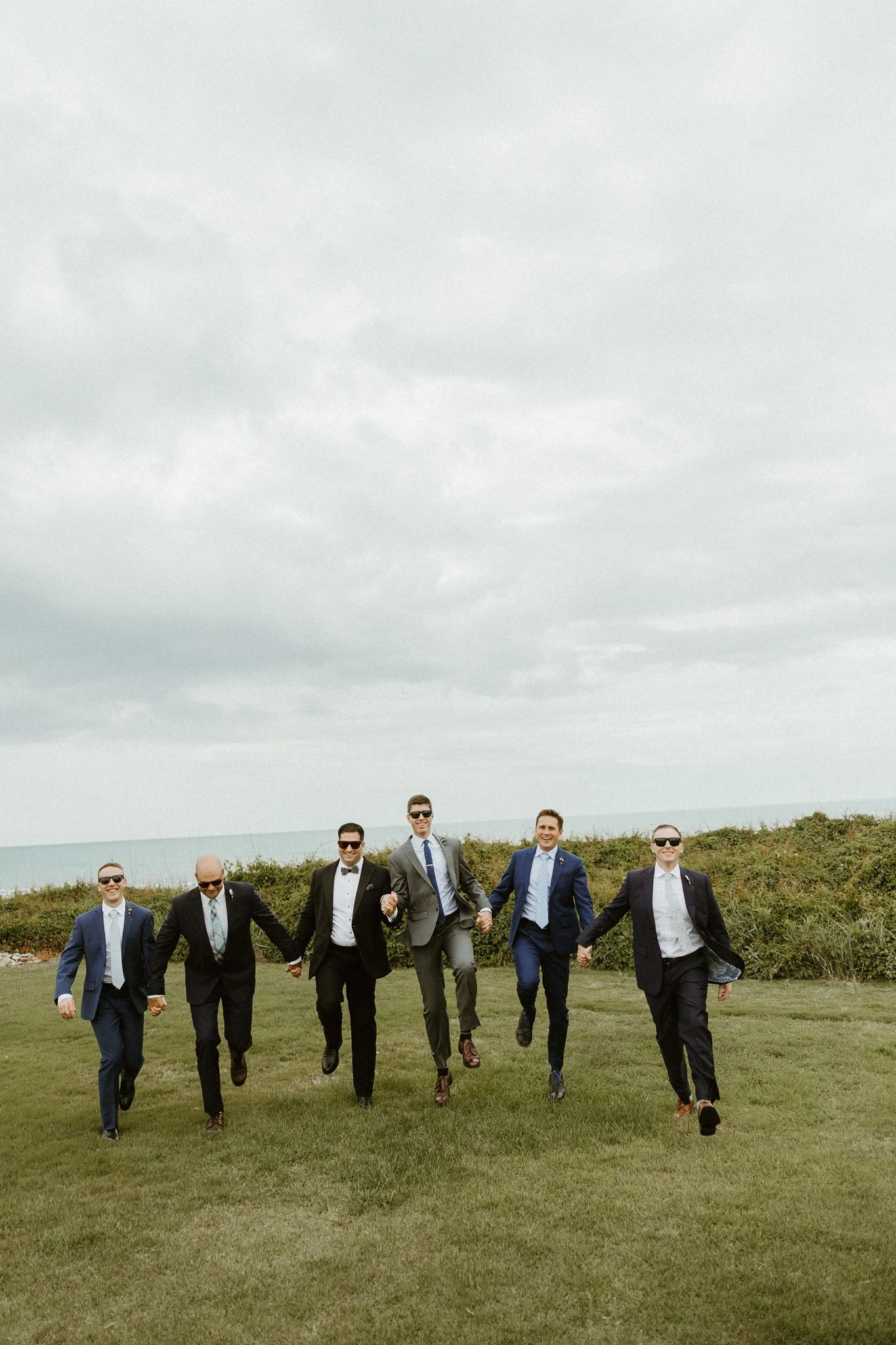 Group of seven men in suits walking on grass outdoors near the ocean under a cloudy sky.