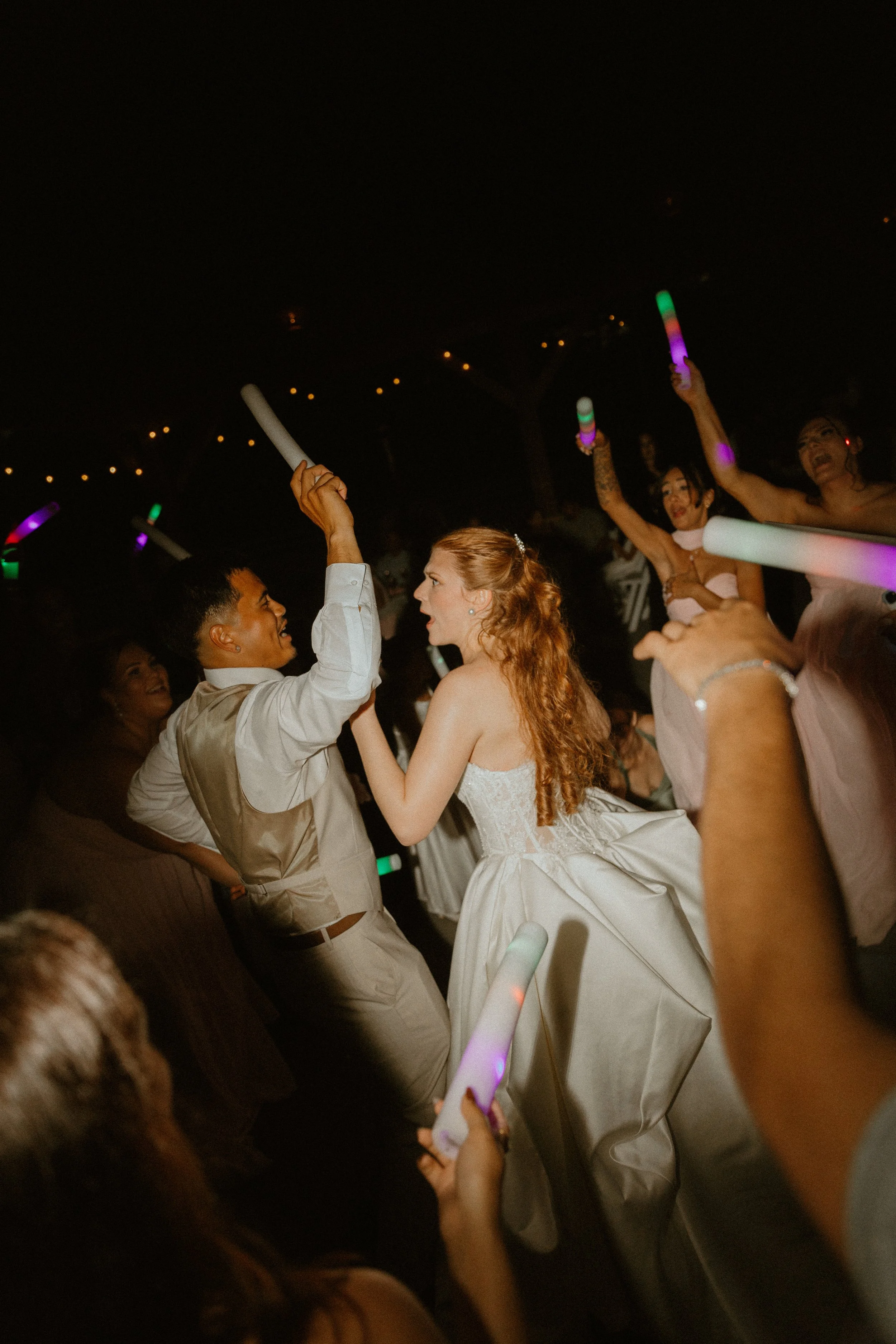 People dancing at a nighttime party, holding glow sticks and wearing formal attire, including a woman in a white dress and a man in a beige suit.
