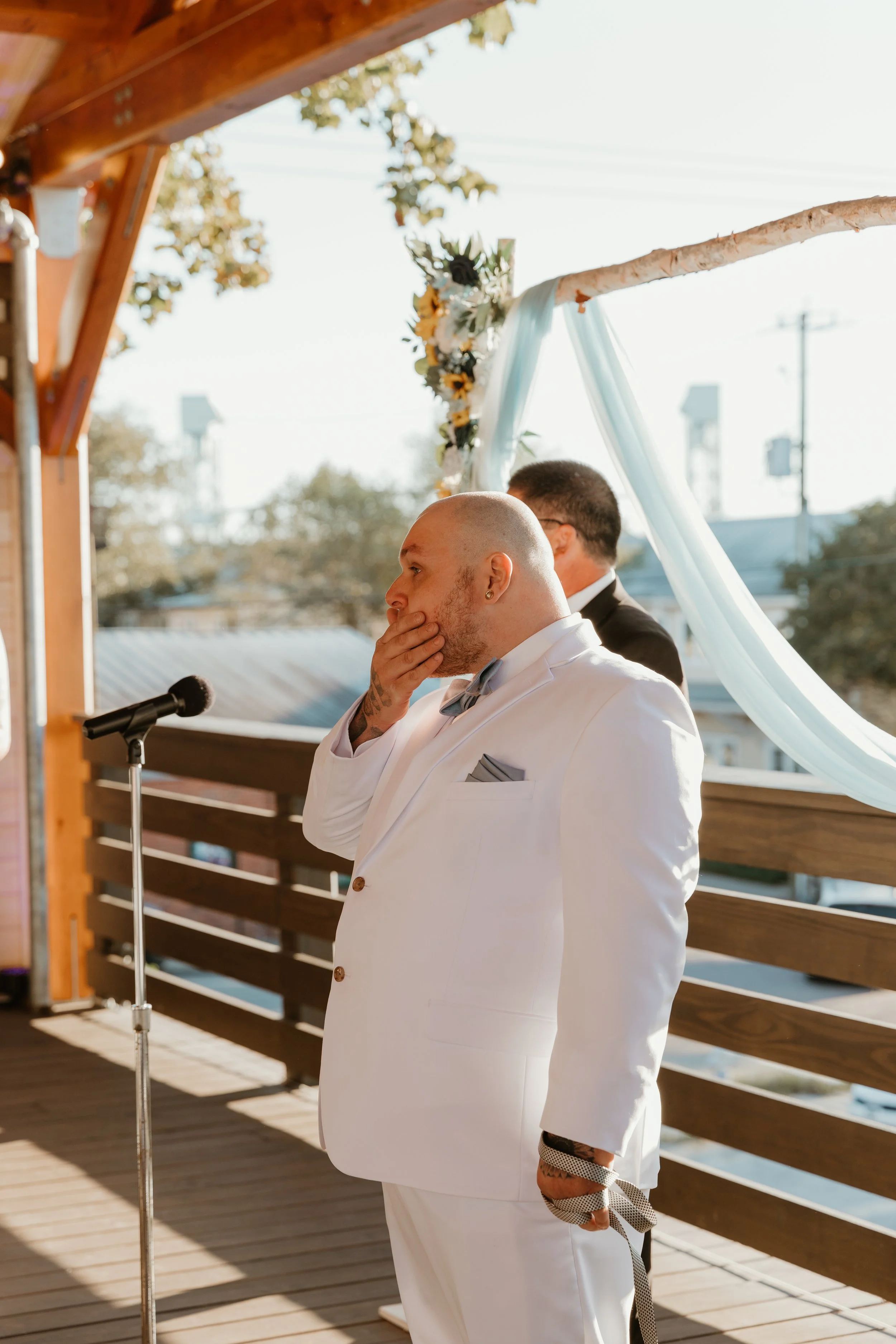 A man in a white suit with a bow tie standing in front of a microphone on an outdoor wooden deck, with another man in a black suit behind him, during a wedding ceremony.