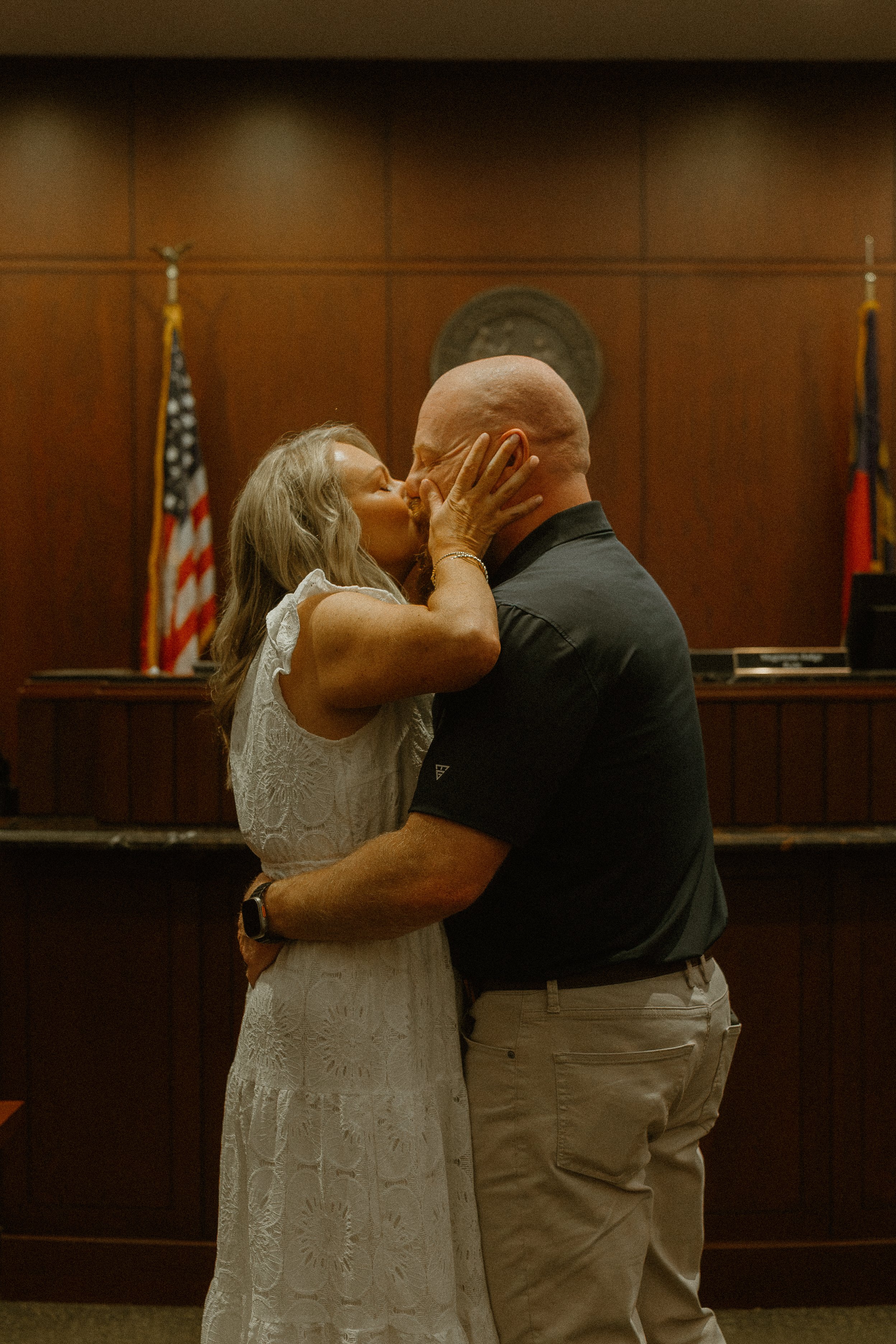 An older woman with long gray hair wearing a white lace dress kissing a bald man with a beard dressed in dark clothing. They are in a formal room with American and another flag in the background facing each other, and a wooden panel wall.