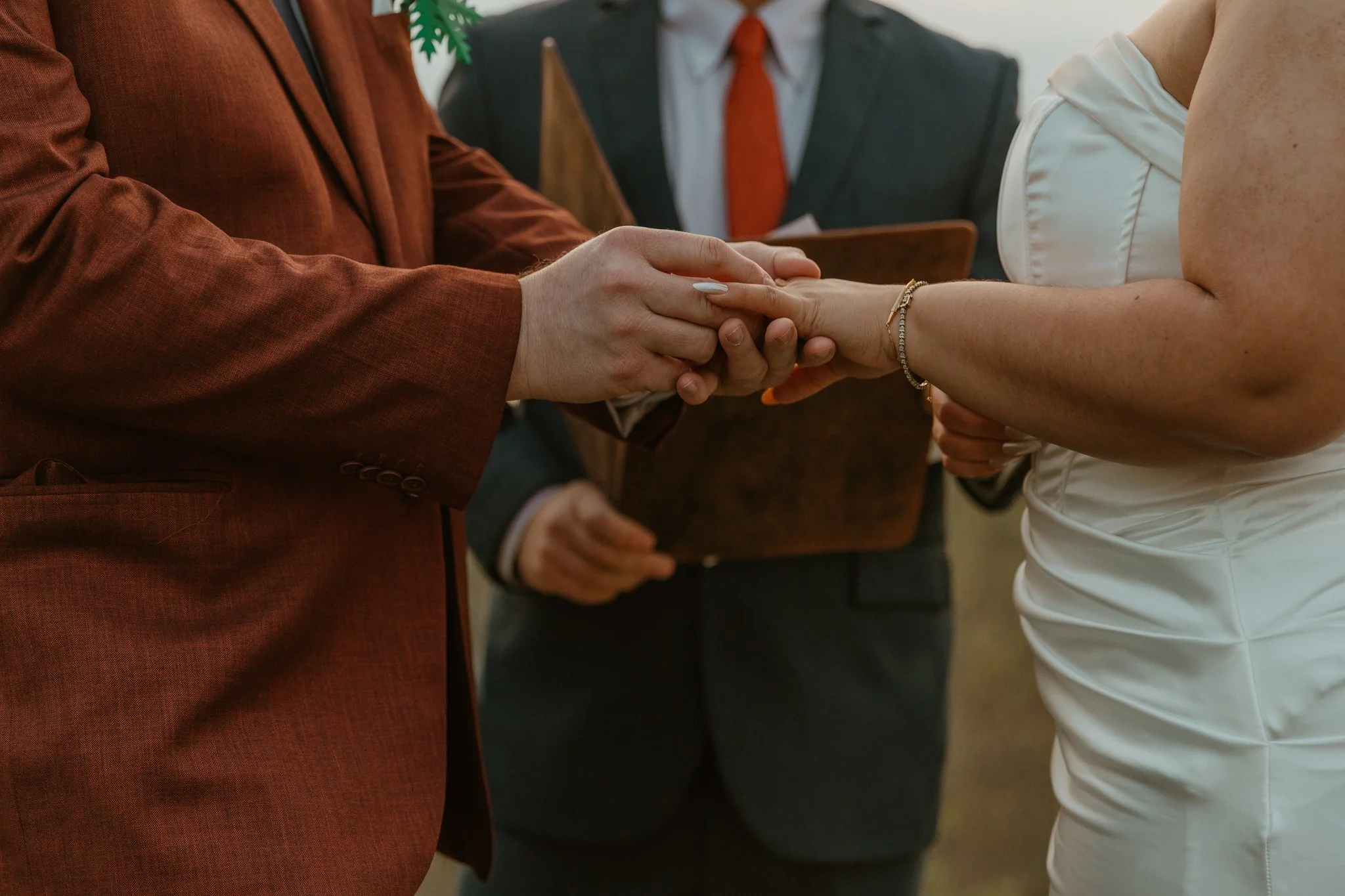 Two people holding hands during a wedding ceremony, with a person in a suit and tie holding a tablet in the background.