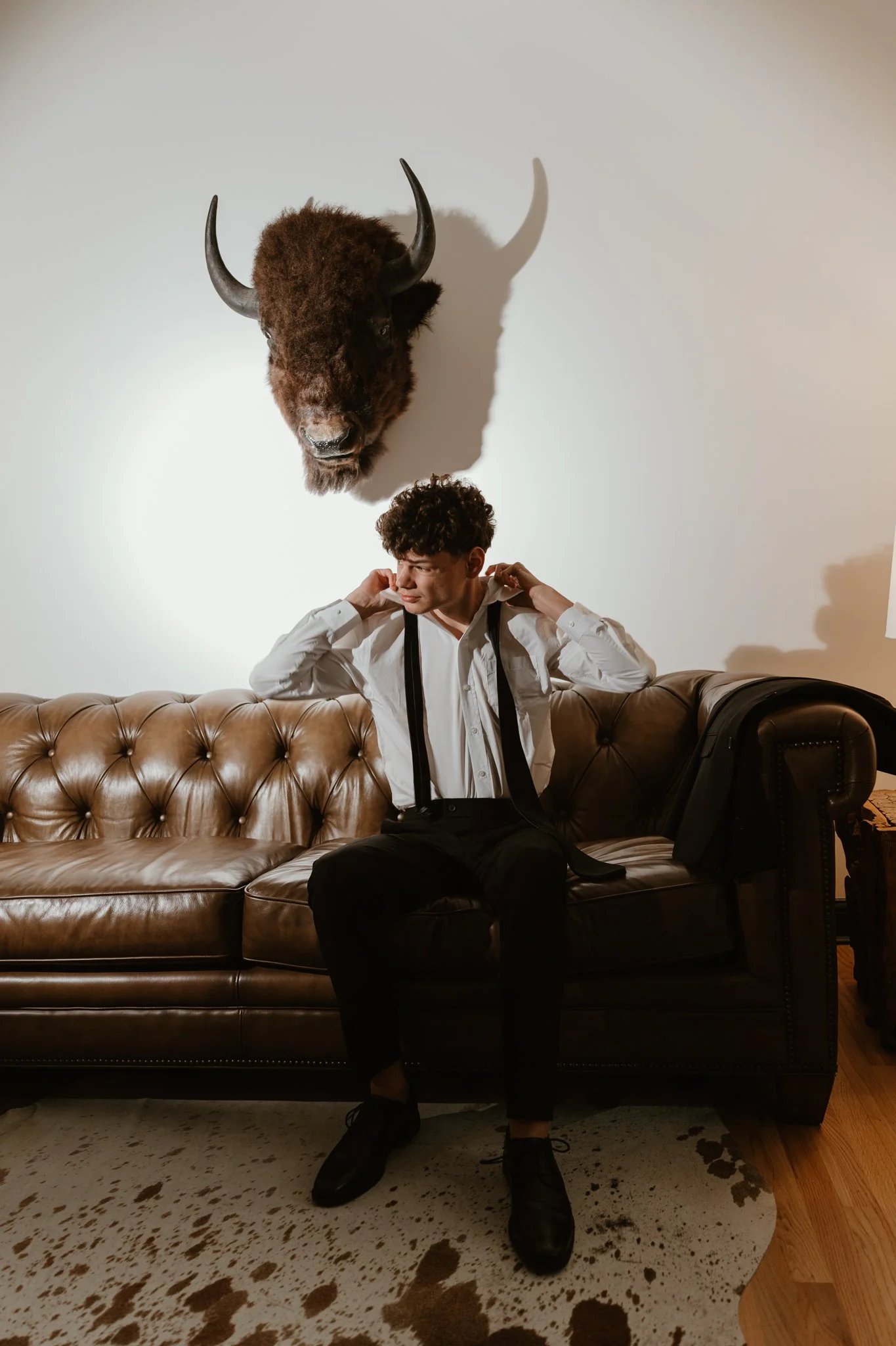 A young man in formal attire sitting on a brown leather couch, adjusting his suspenders, with a bison head mounted on the wall behind him and a backpack on the floor beside him.