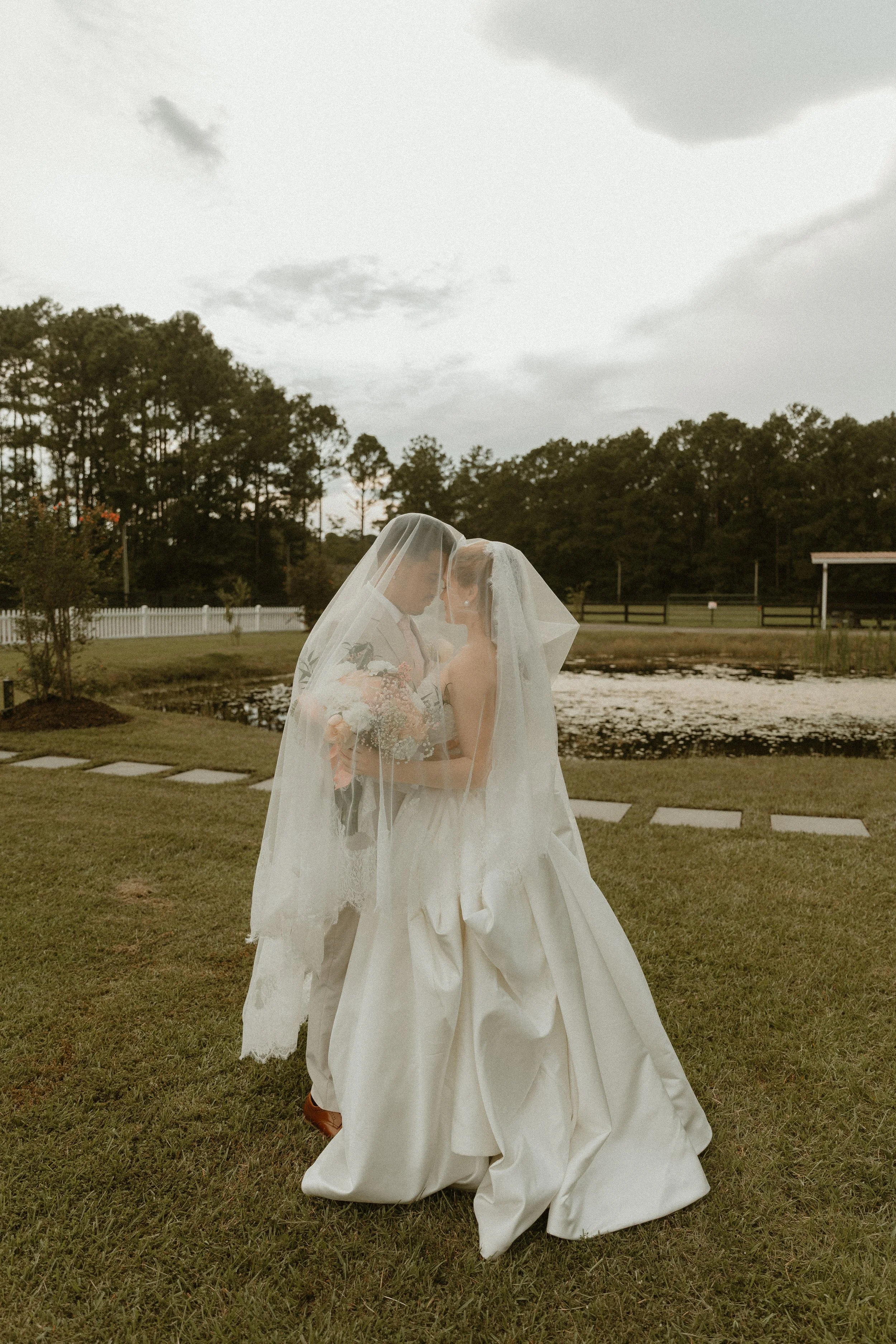 A bride and groom standing closely together outdoors, sharing a kiss behind a wedding veil, with a pond, trees, and a gazebo in the background, and cloudy sky overhead.