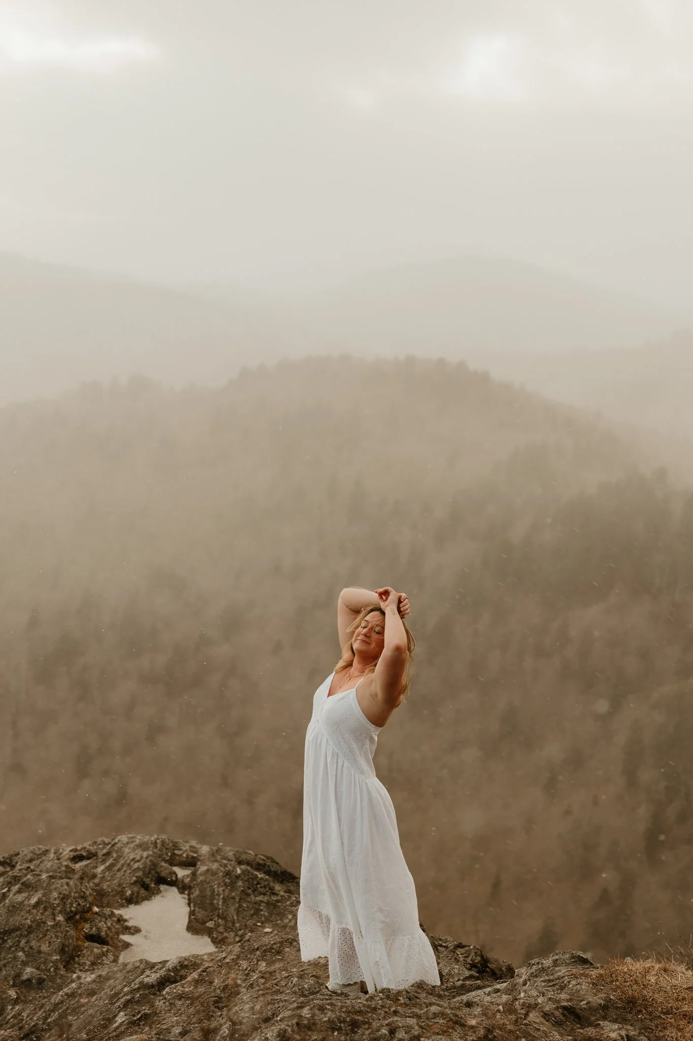Woman in a white dress standing on a rocky landscape in front of a misty, mountainous background during rain.