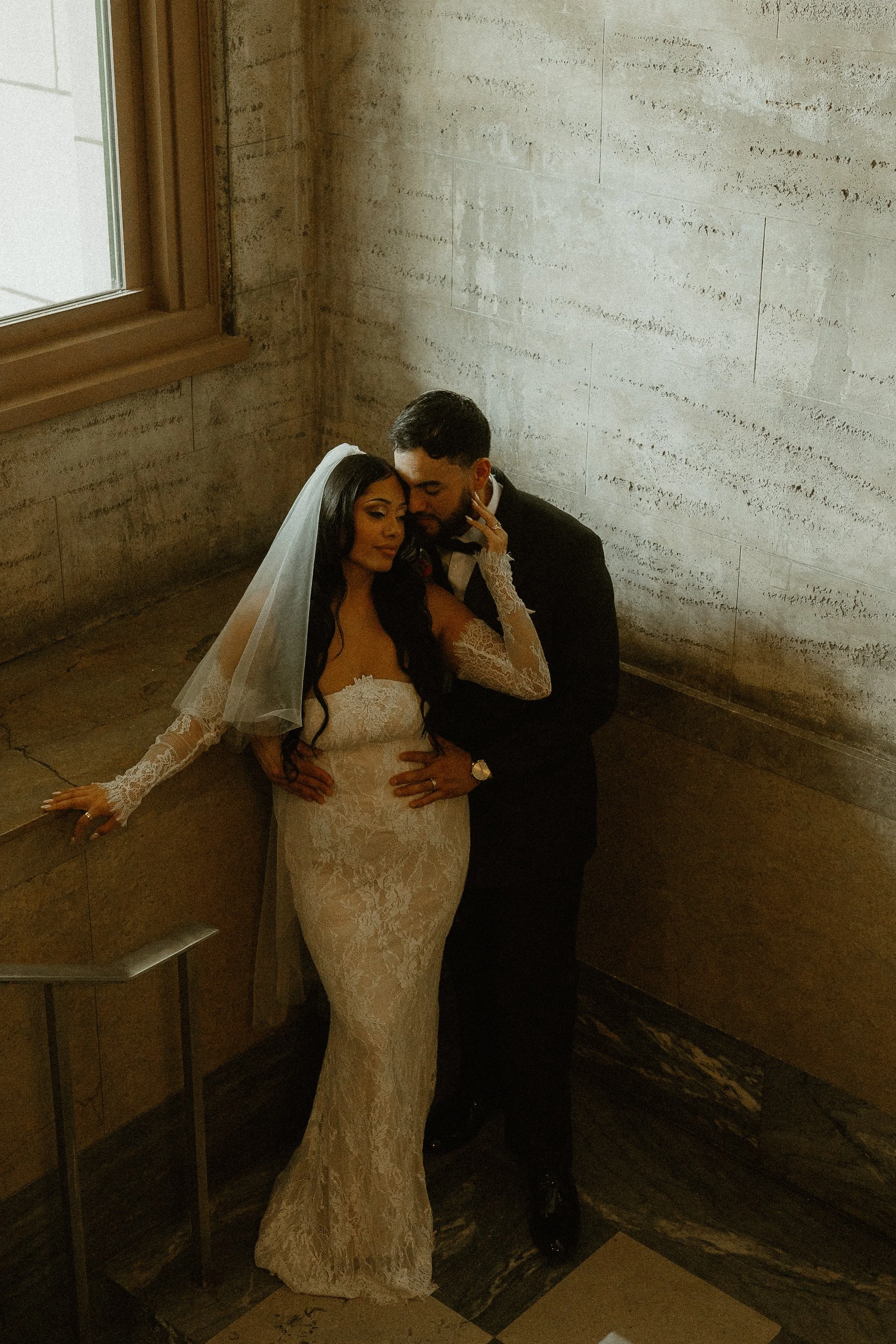 A bride and groom embracing in a dimly lit, rustic indoor setting with a concrete wall and a window nearby.