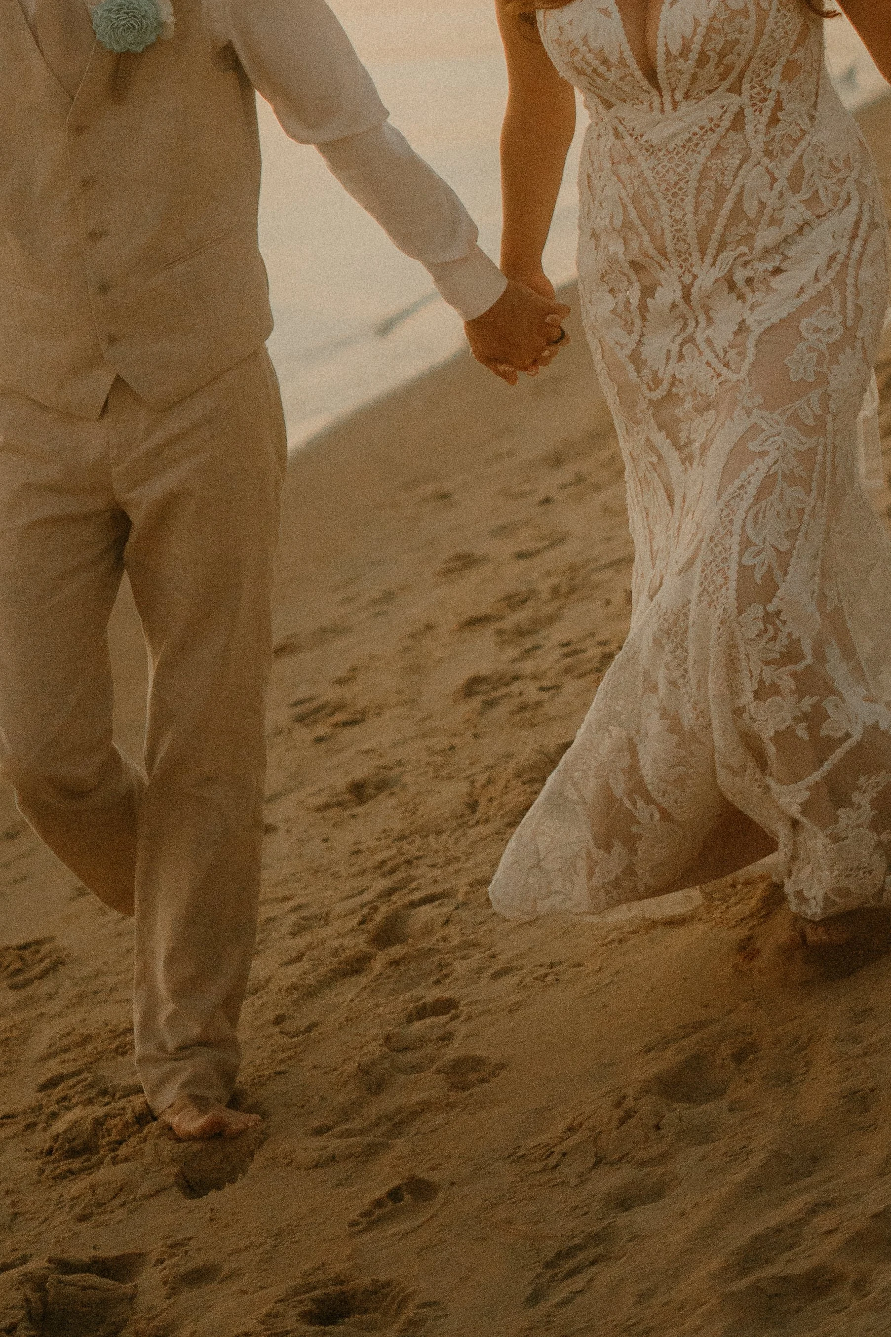 A couple holding hands while walking barefoot on a sandy beach, with the woman wearing a white lace dress and the man dressed in light-colored trousers and a vest.