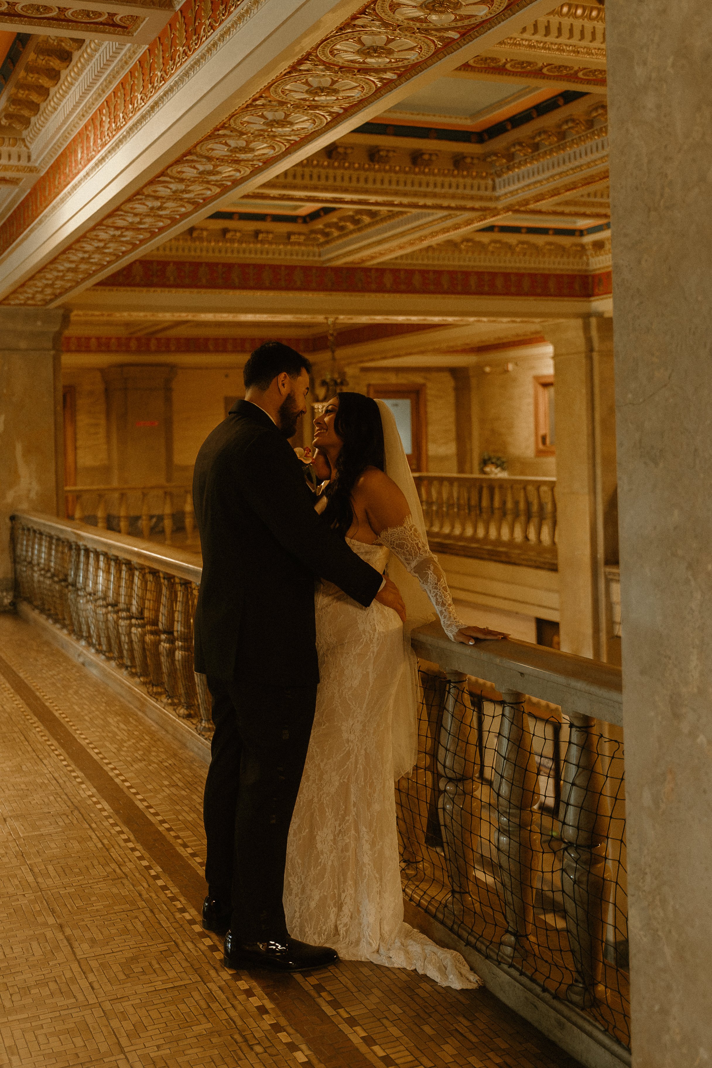 A bride and groom sharing a moment on a balcony in an ornate, historic building. The groom is dressed in a black tuxedo, and the bride in a white lace wedding gown and veil. They are smiling and looking at each other, with the bride resting her hand 
