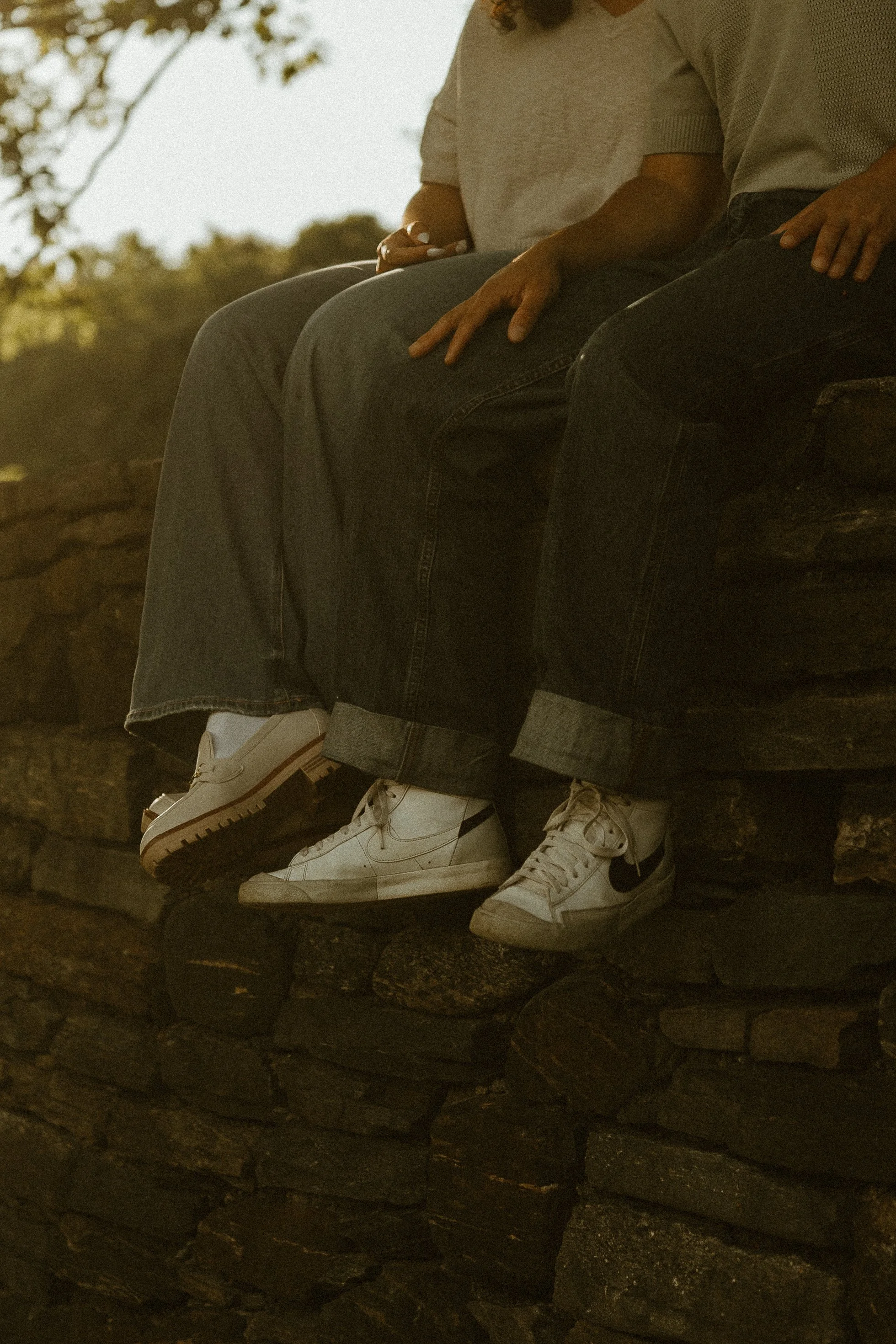 Close-up of two people sitting on a stone ledge, showing their legs and shoes, with a blurred outdoor background.