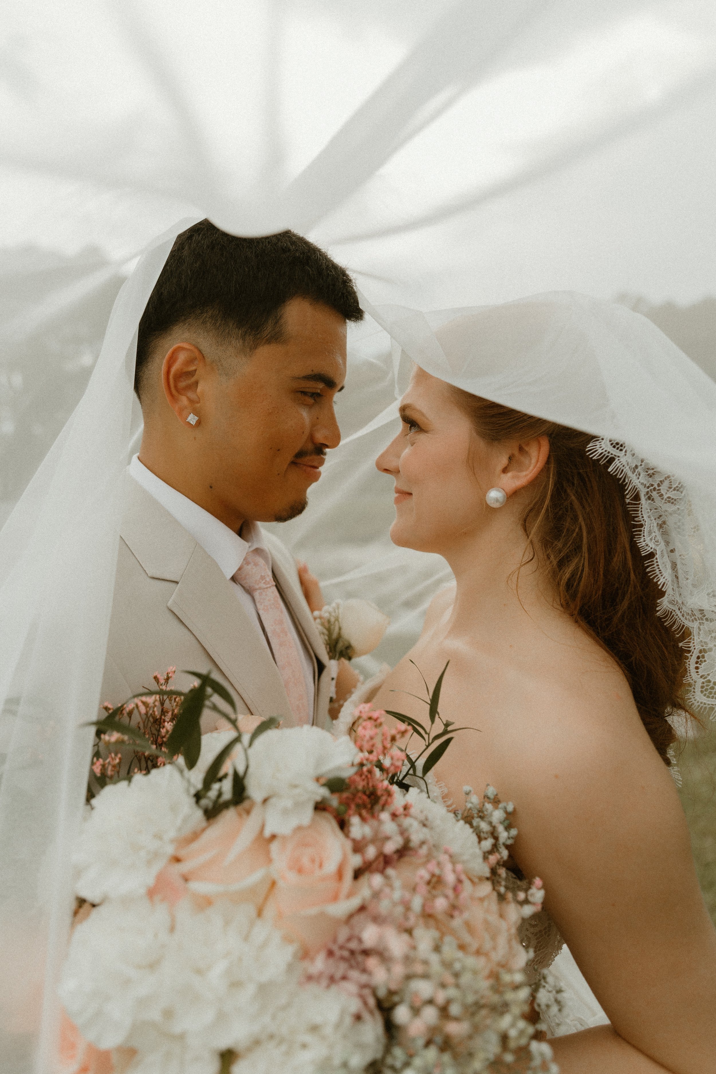 A bride and groom face each other under a sheer white veil, with the bride holding a bouquet of pink and white flowers.