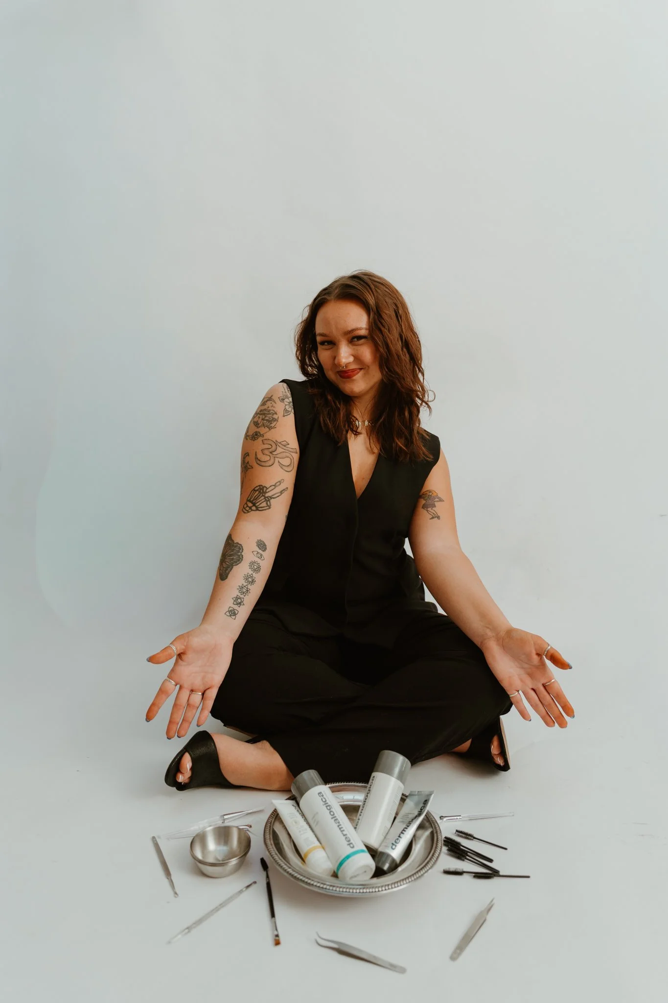 A woman with tattoos sitting cross-legged on the floor, smiling, with skincare products and tools arranged in a circle around her, against a plain white background.