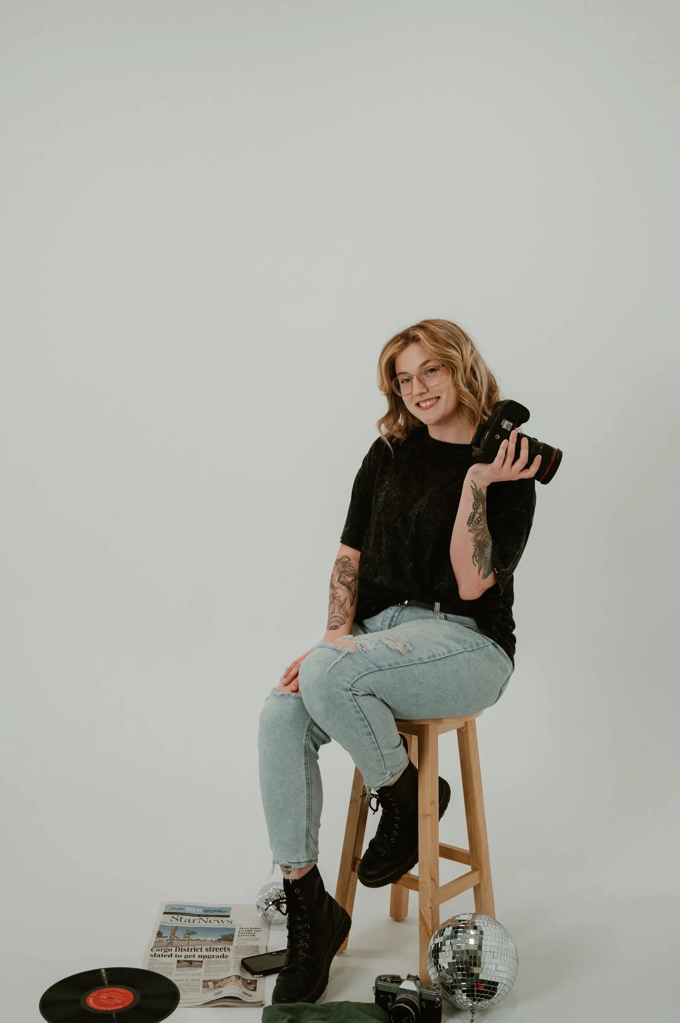 A woman with glasses and tattoos, smiling while holding a camera, sitting on a wooden stool in front of a plain white background. There are items on the floor including a vinyl record, a newspaper, a camera, a disco ball, and a pair of shoes.
