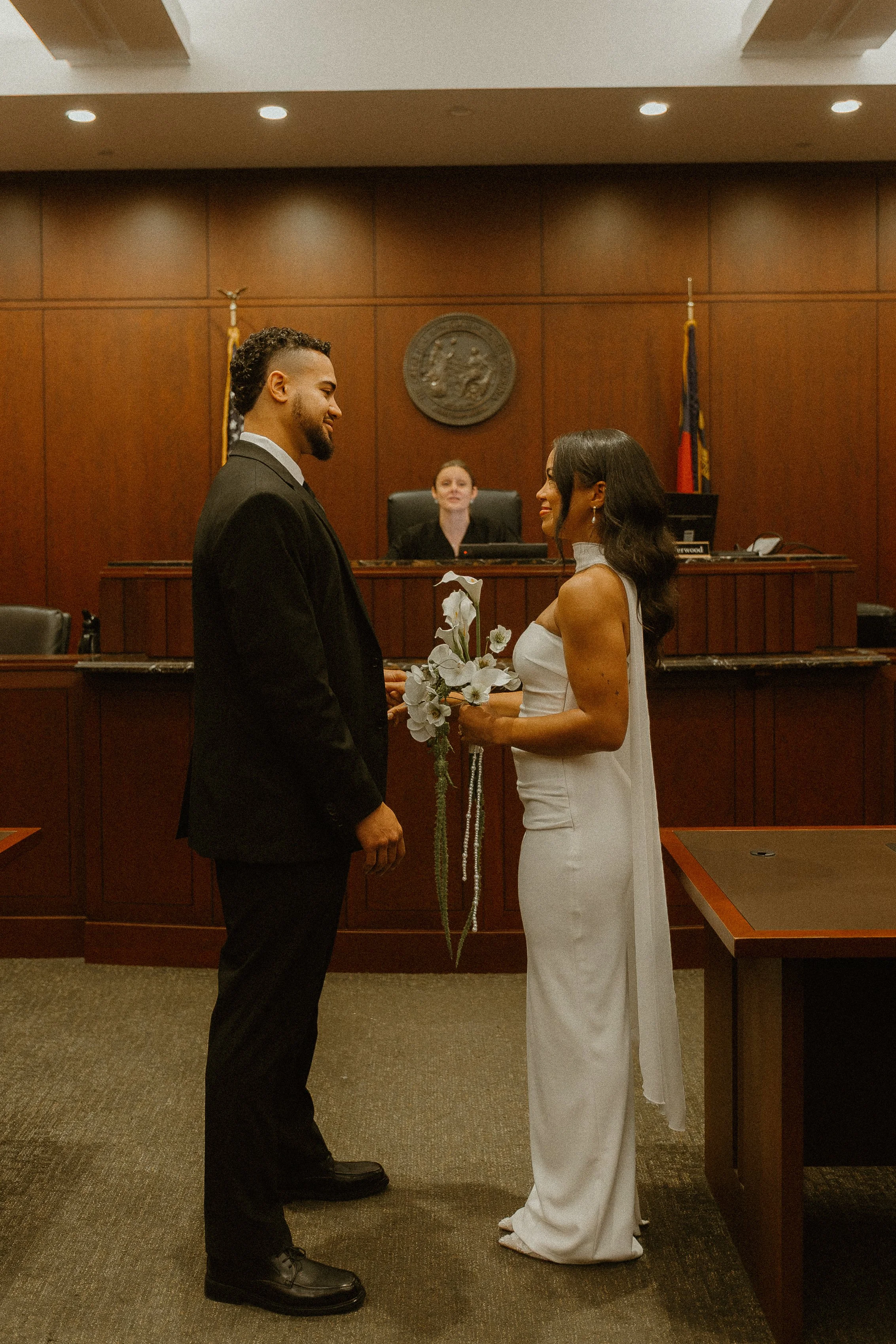 A couple stands face to face in a courtroom, holding a bouquet of white flowers and rosary beads, with a judge seated behind them at the bench.