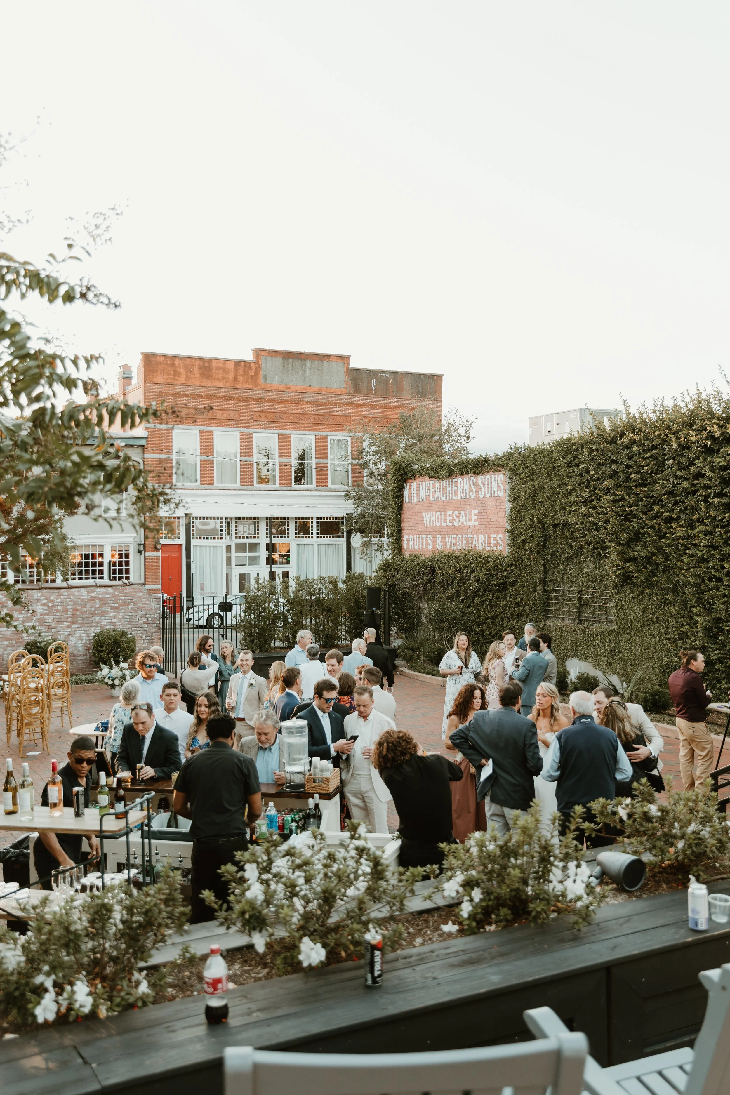 People gathered outdoors at a social event, with a bar setup serving drinks, in front of a brick building and lush greenery