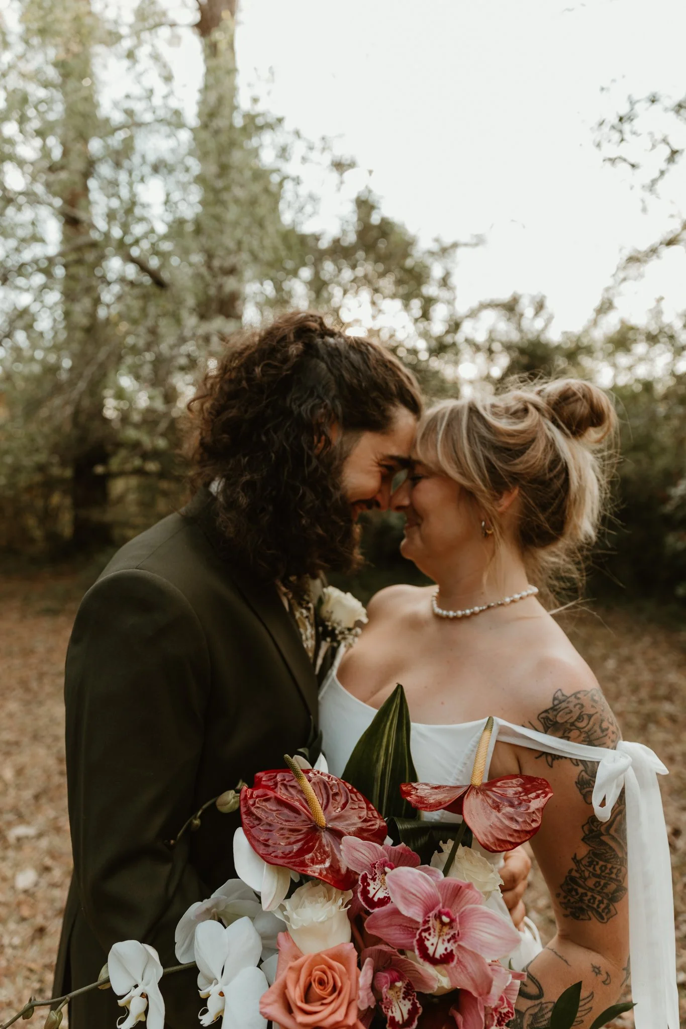 A couple dressed in wedding attire sharing a close, intimate moment outdoors surrounded by trees, with the bride holding a large bouquet of flowers.