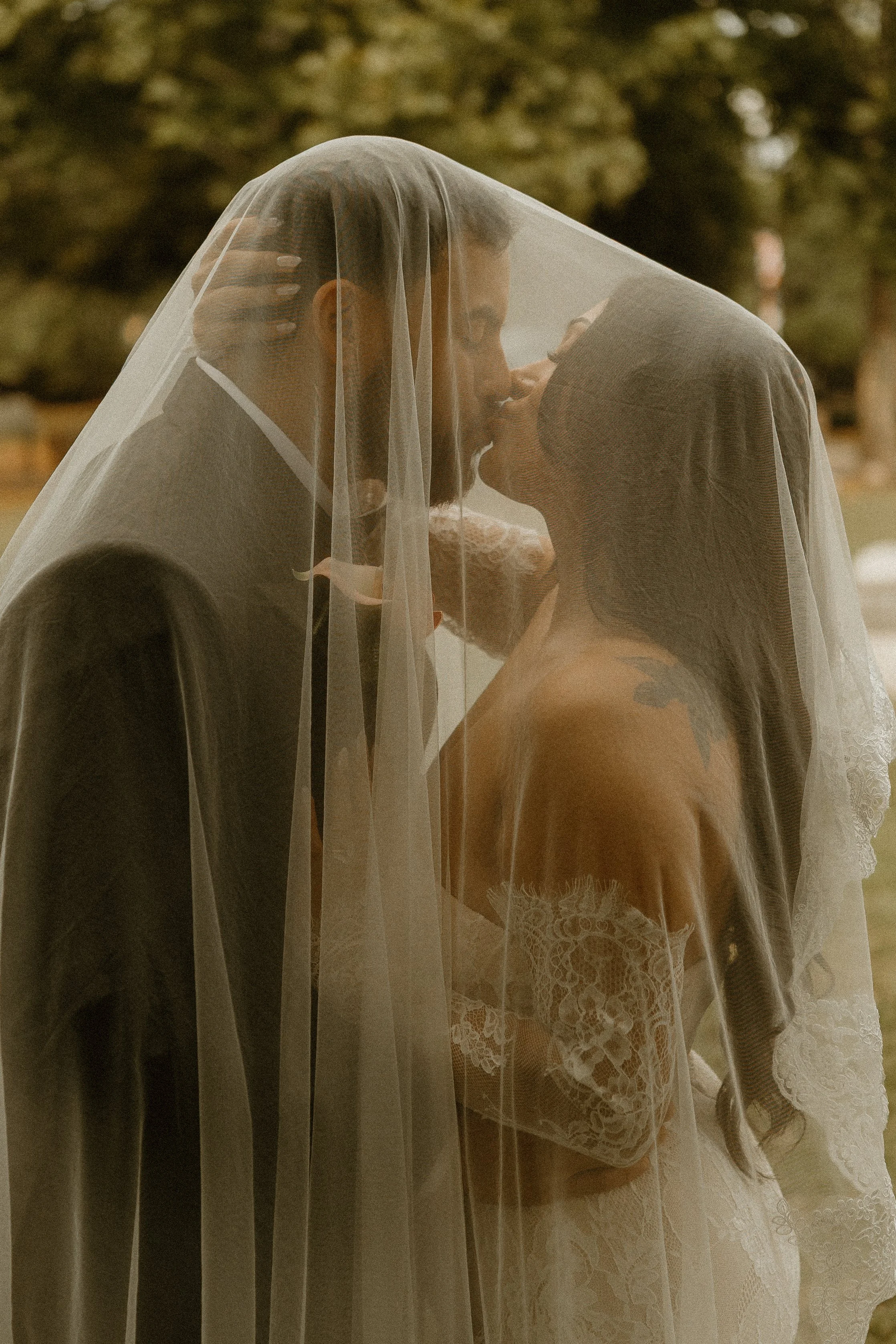 A bride and groom kissing under a veil outdoors, with trees and blurred background.
