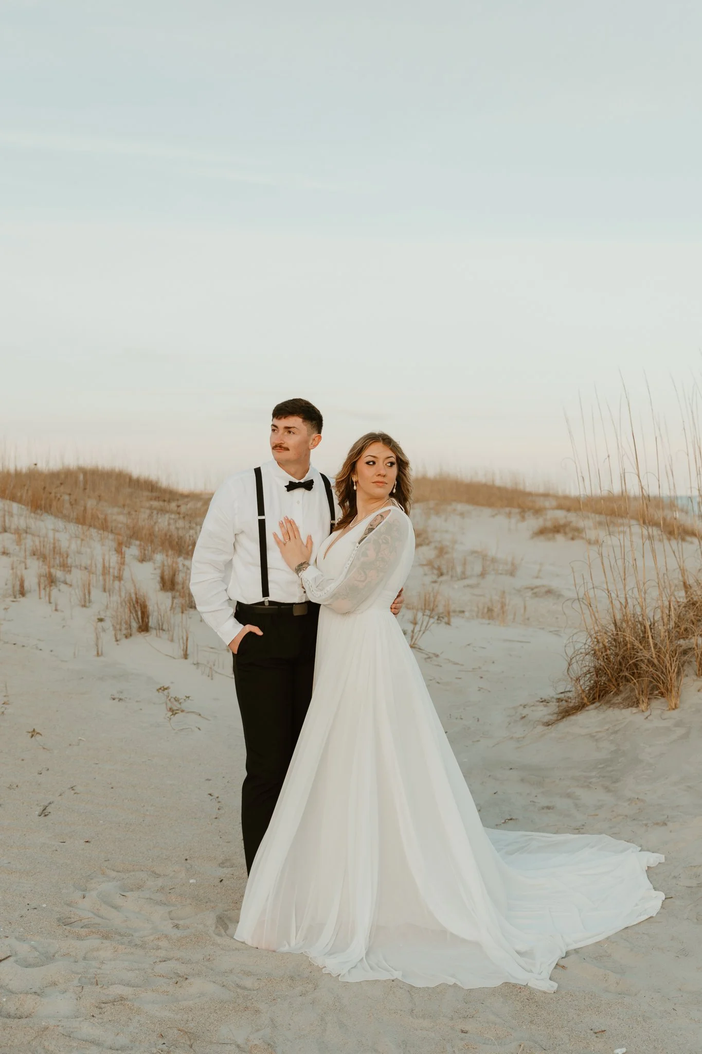 A couple in wedding attire standing on a sandy beach with dry grass, under a clear sky.