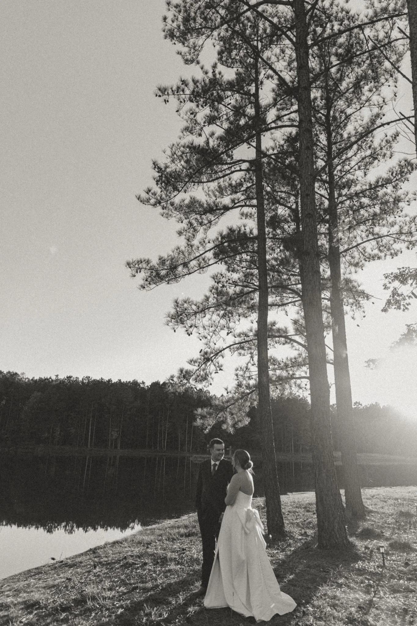 A black and white photo of a couple in formal attire, standing outdoors beside a body of water, with tall trees and a forest in the background, in the late afternoon sunlight.