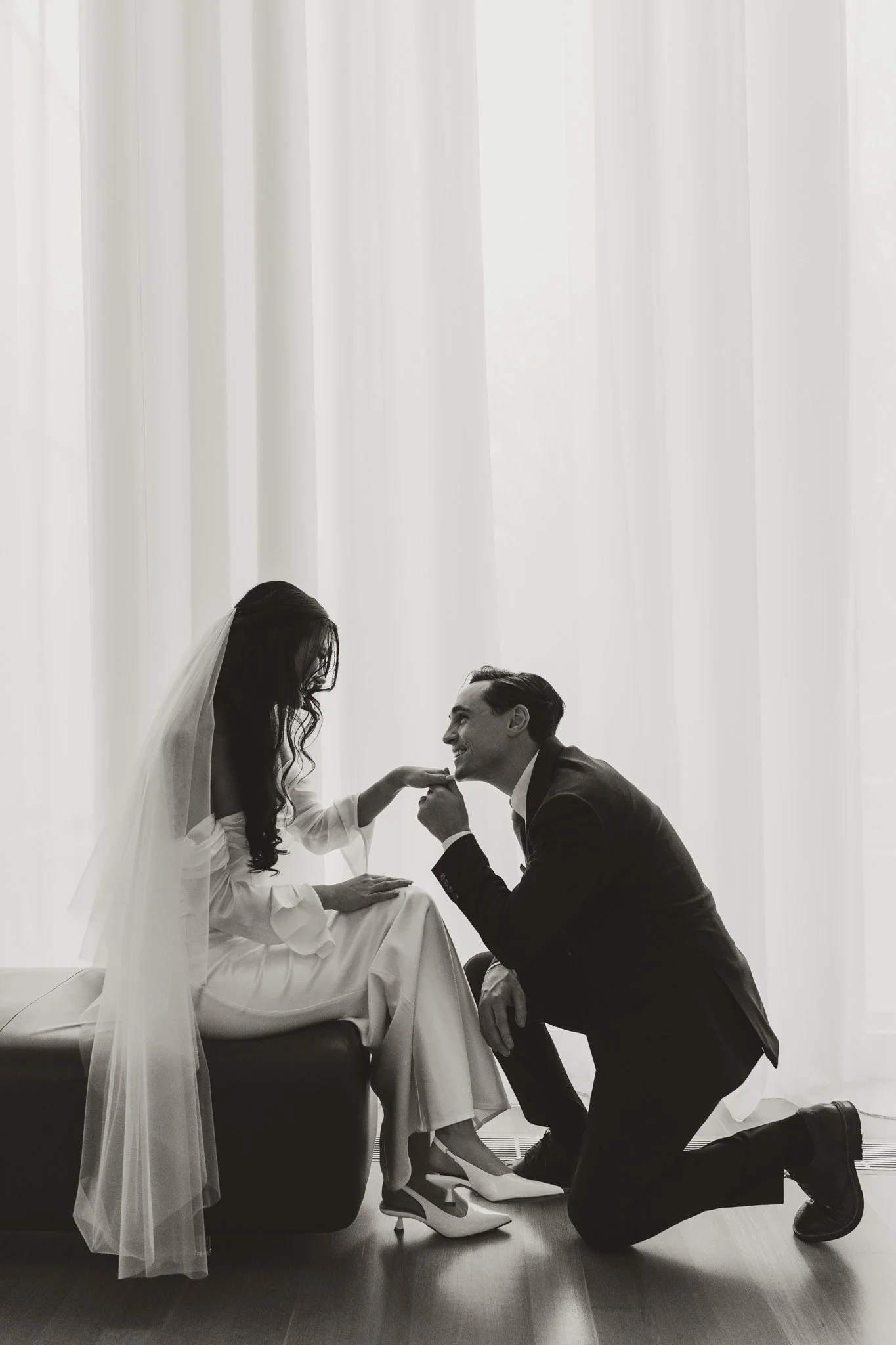 A black and white photo of a bride and groom. The groom is kneeling and holding the bride's hand, and they are looking into each other's eyes. The bride is sitting on a bench wearing a wedding dress and veil, and the groom is in a suit. They are in f