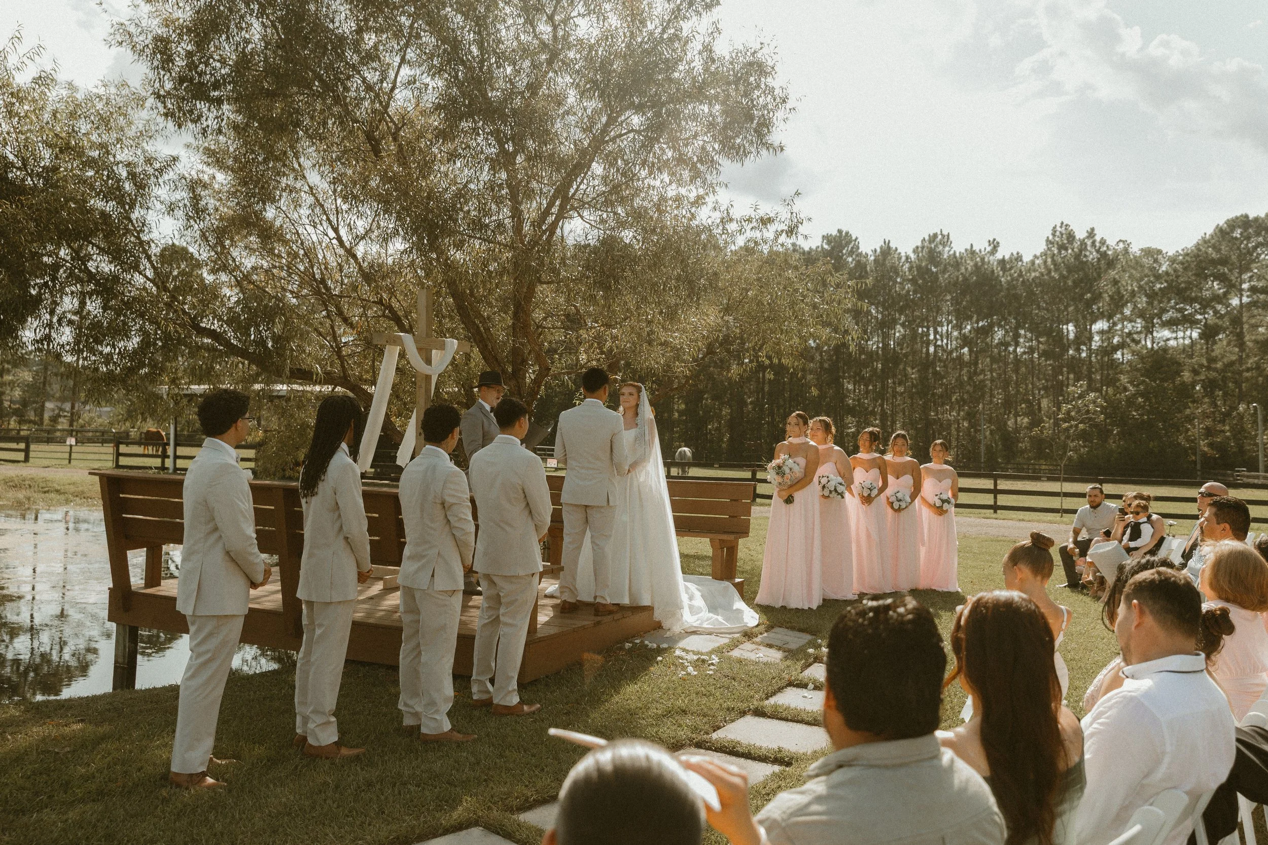 A wedding ceremony taking place outdoors on a wooden platform by a pond, with a bride and groom exchanging vows. Bridesmaids in pink dresses and groomsmen in light suits are present, and guests are seated watching the event.